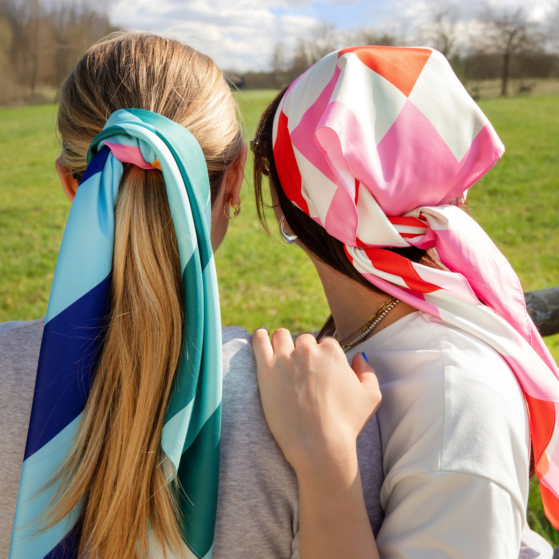 Twee vrouwen van achteren, de ene met een sjaal als haarband, de andere met een sjaal als hoofddoek. Een weiland en bomen op de achtergrond.