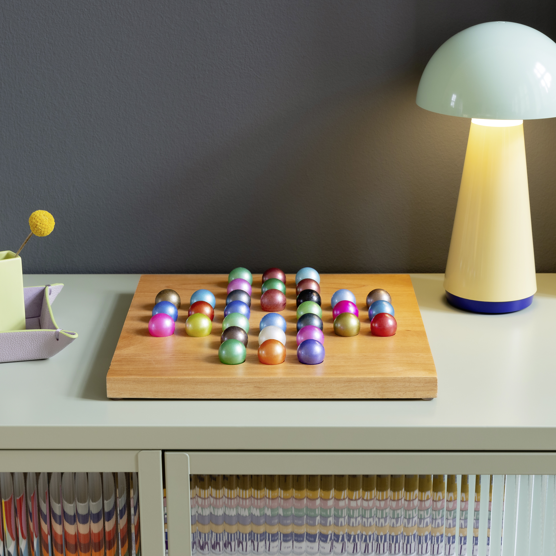 Wooden board with 32 coloured glass balls arranged in rows, as a solitaire game on a light green sideboard with a yellow lamp.