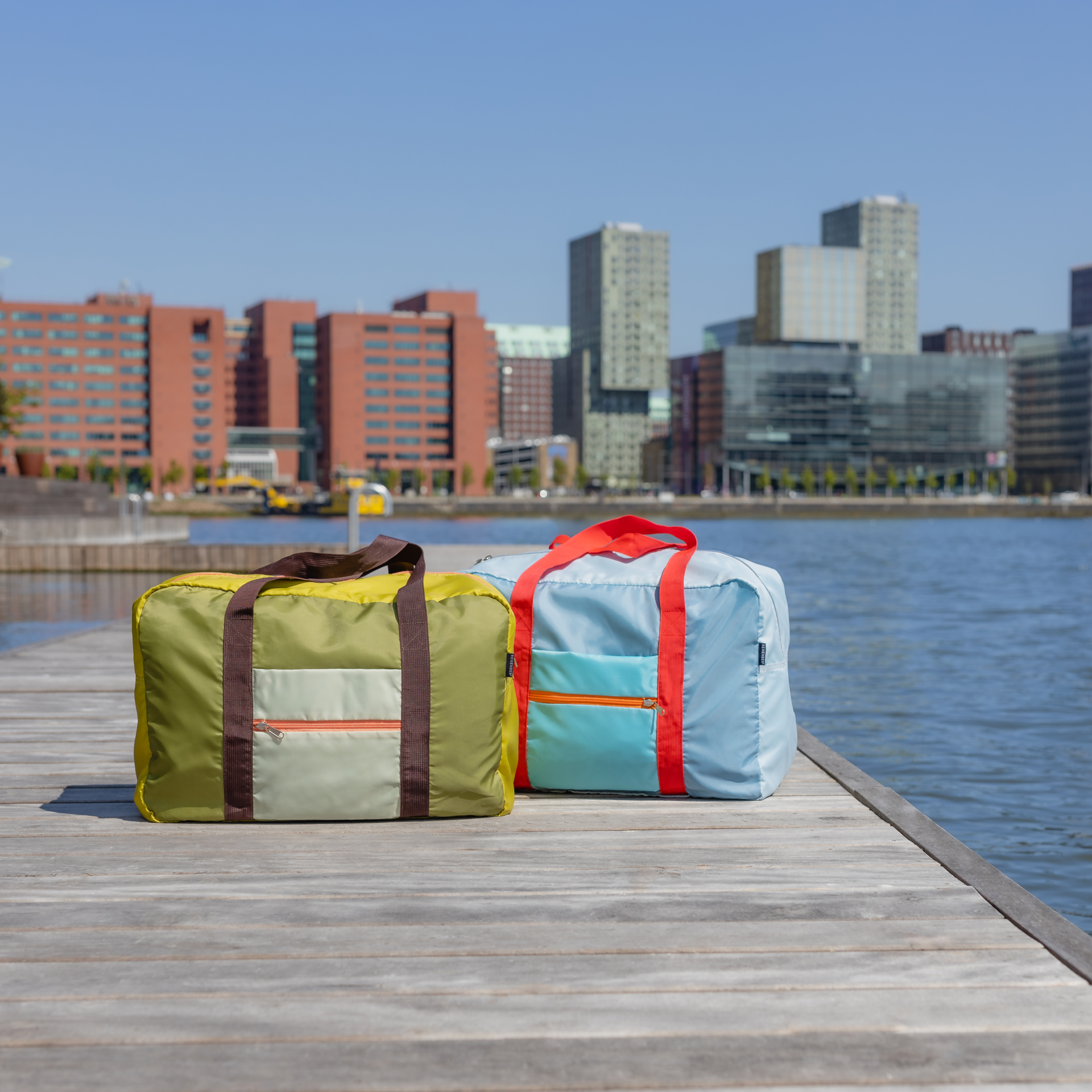 Dos bolsas de viaje «Liz», una verde claro y otra azul claro, descansan sobre una pasarela de madera frente a un horizonte con rascacielos y aguas azules. Las bolsas tienen asas a juego.