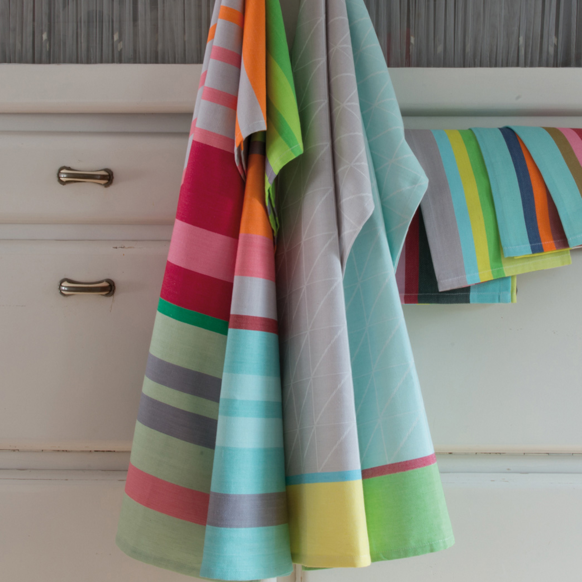 Plusieurs Set de torchons n° 16 sont suspendus à une commode blanche. Les chiffons sont rayés et ornés de motifs géométriques aux couleurs vives.