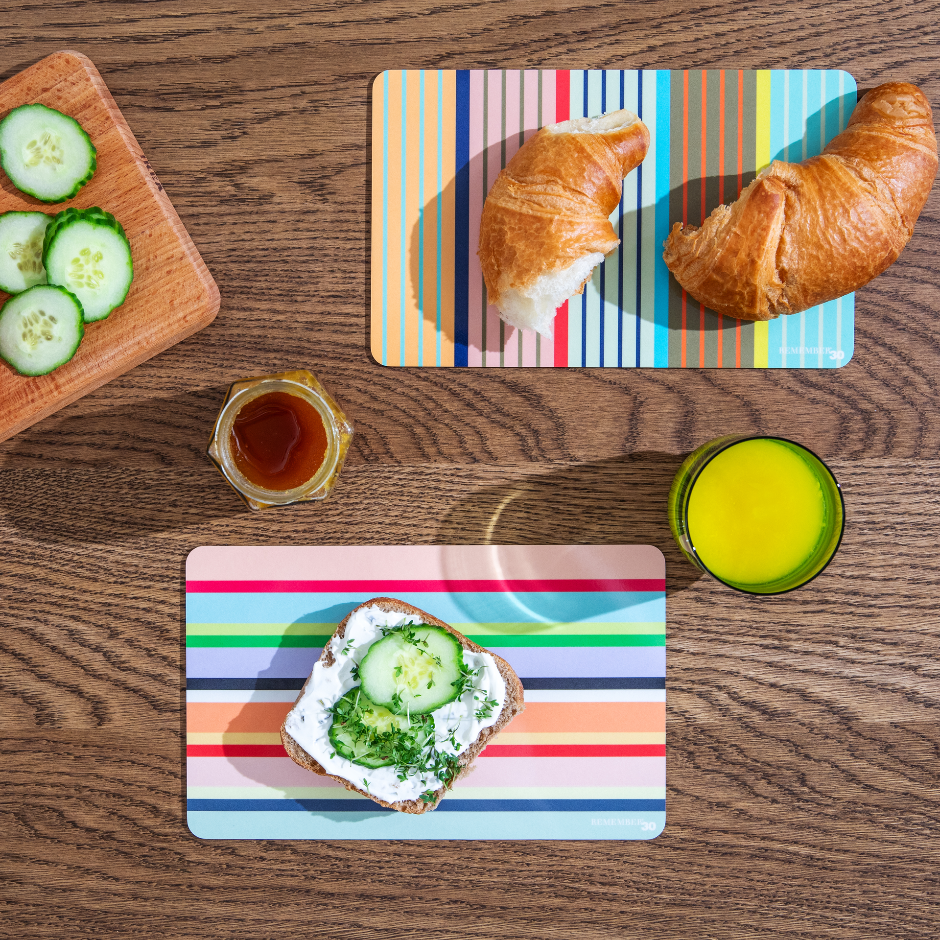 Draufsicht: Frühstücksbrettchen Stripes mit Croissant, Brot mit Frischkäse und Gurke, Honig und Saft auf Holztisch.