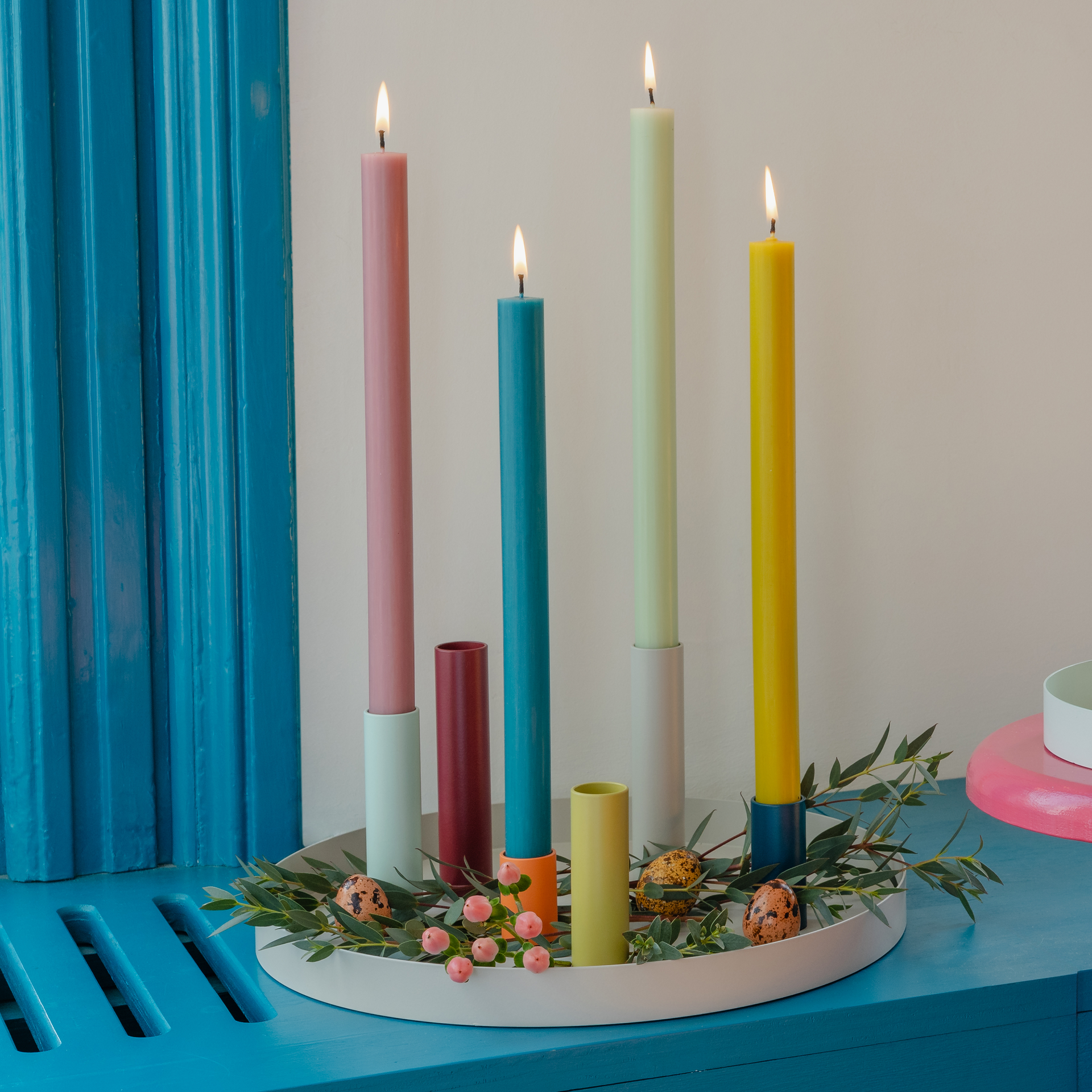 Una composizione di candele colorate e portacandele coordinati su un vassoio rotondo bianco. Decorata con elementi floreali e uova decorative. Alcune candele sono accese.