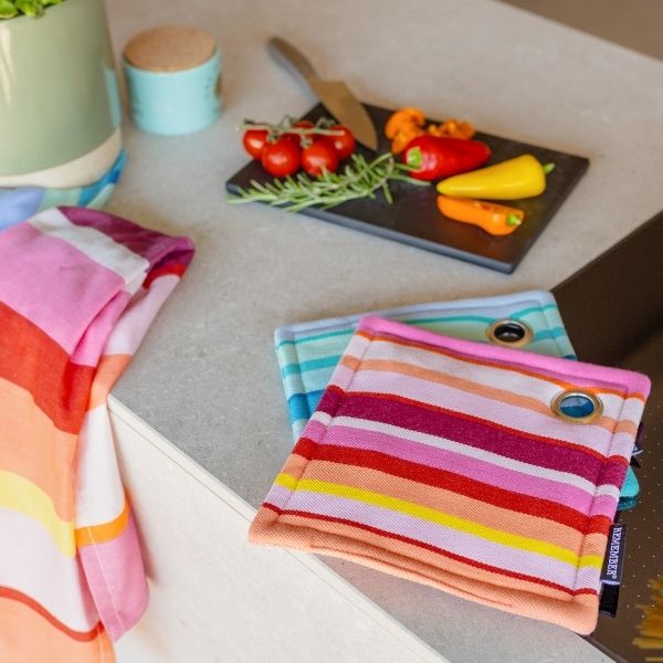 Zwei bunte, gestreifte Topflappen und ein passendes Küchentuch liegen auf einer Küchenarbeitsplatte. Auf einem schwarzen Schneidebrett im Hintergrund: Messer, Tomaten, Rosmarin und Paprika. Eine grüne Pflanze rundet ab.