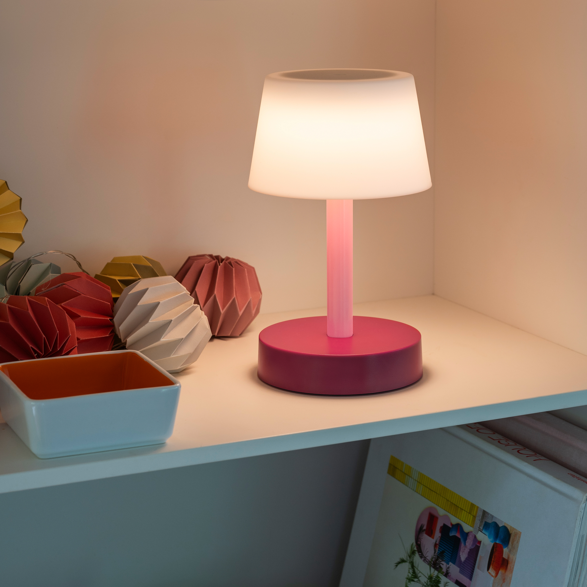 Die Tischleuchte Fritz 'coral' mit eingeschaltetem, warmweißem Licht steht auf einem weißen Regal. Dekorative Faltobjekte und eine Schale stehen daneben.