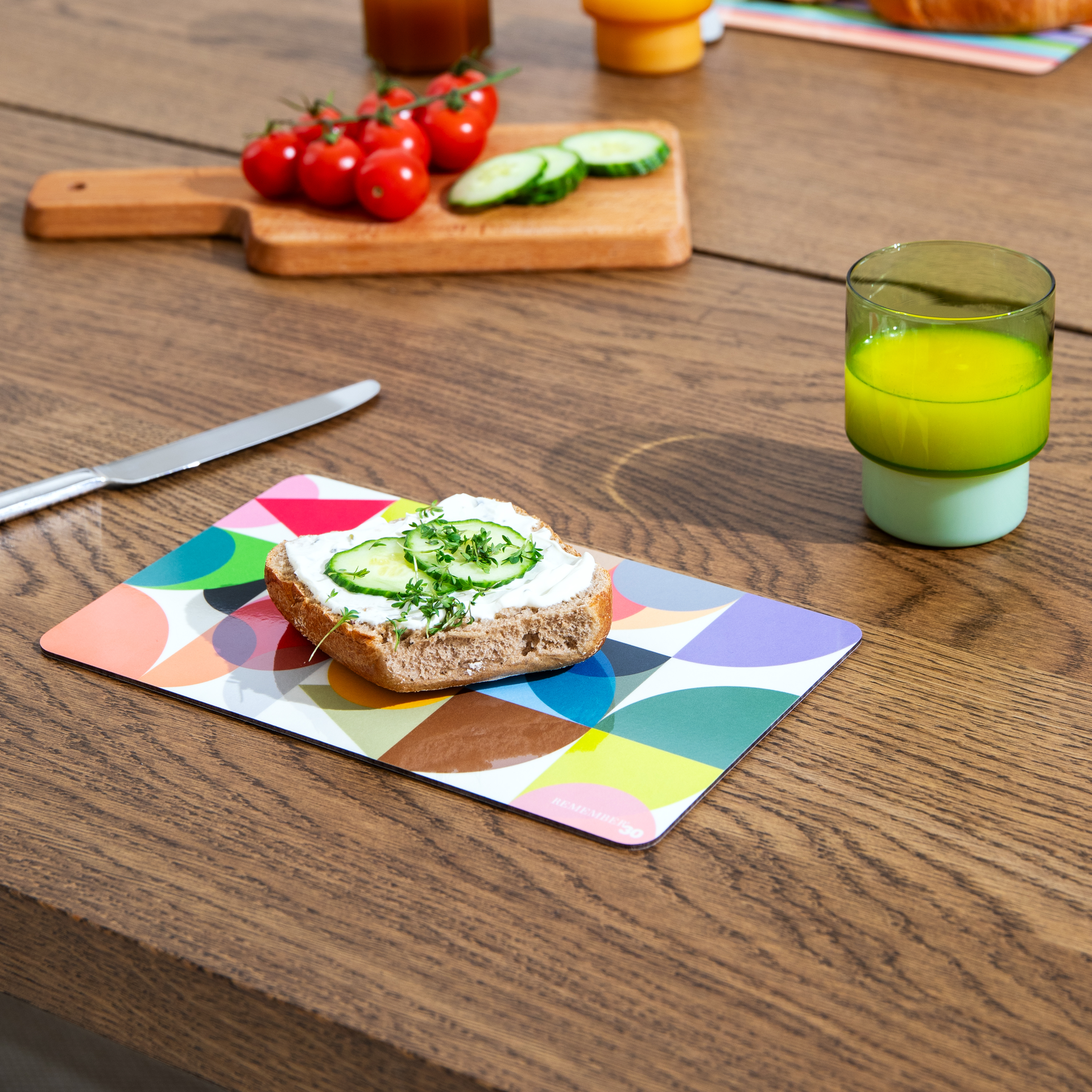 Ein Frühstücksbrettchen 'Remember 30' mit bunten Kreismuster, belegt mit Brot, Frischkäse, Gurke und Kresse. Daneben ein Messer, Tomaten und ein Glas Saft auf einem Holztisch.