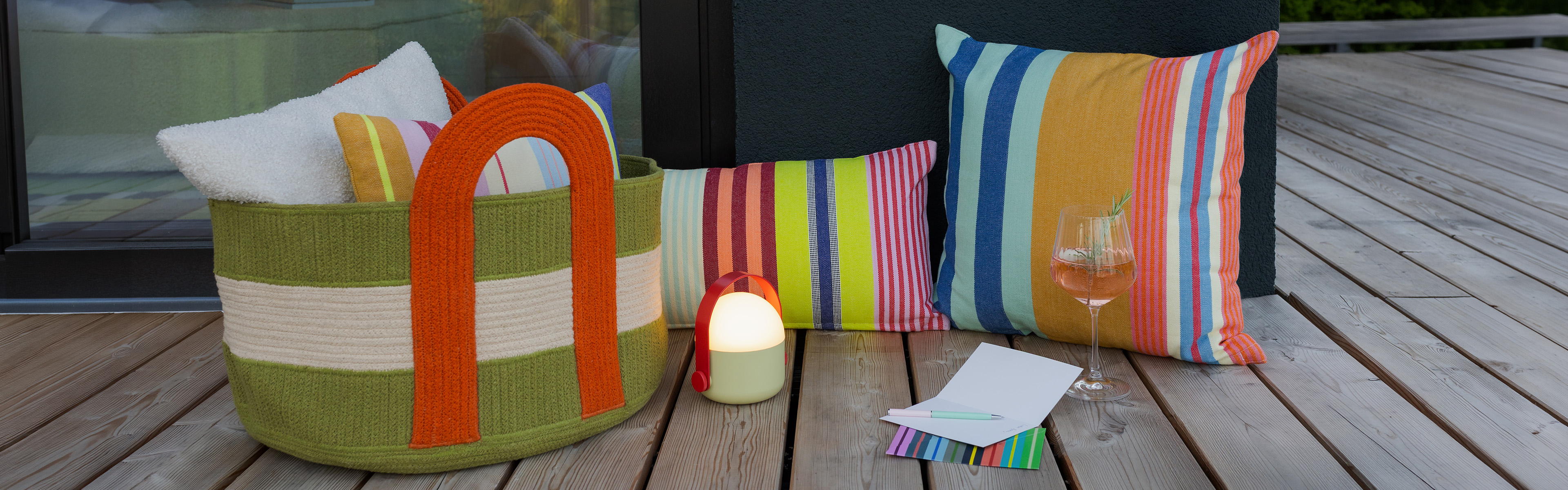 Un espace extérieur chaleureux avec un parquet en bois. À gauche, un grand panier tressé vert et beige aux anses orange, rempli de serviettes blanches et de coussins rayés. À côté, une lampe vert clair. À droite, un verre de vin rosé.