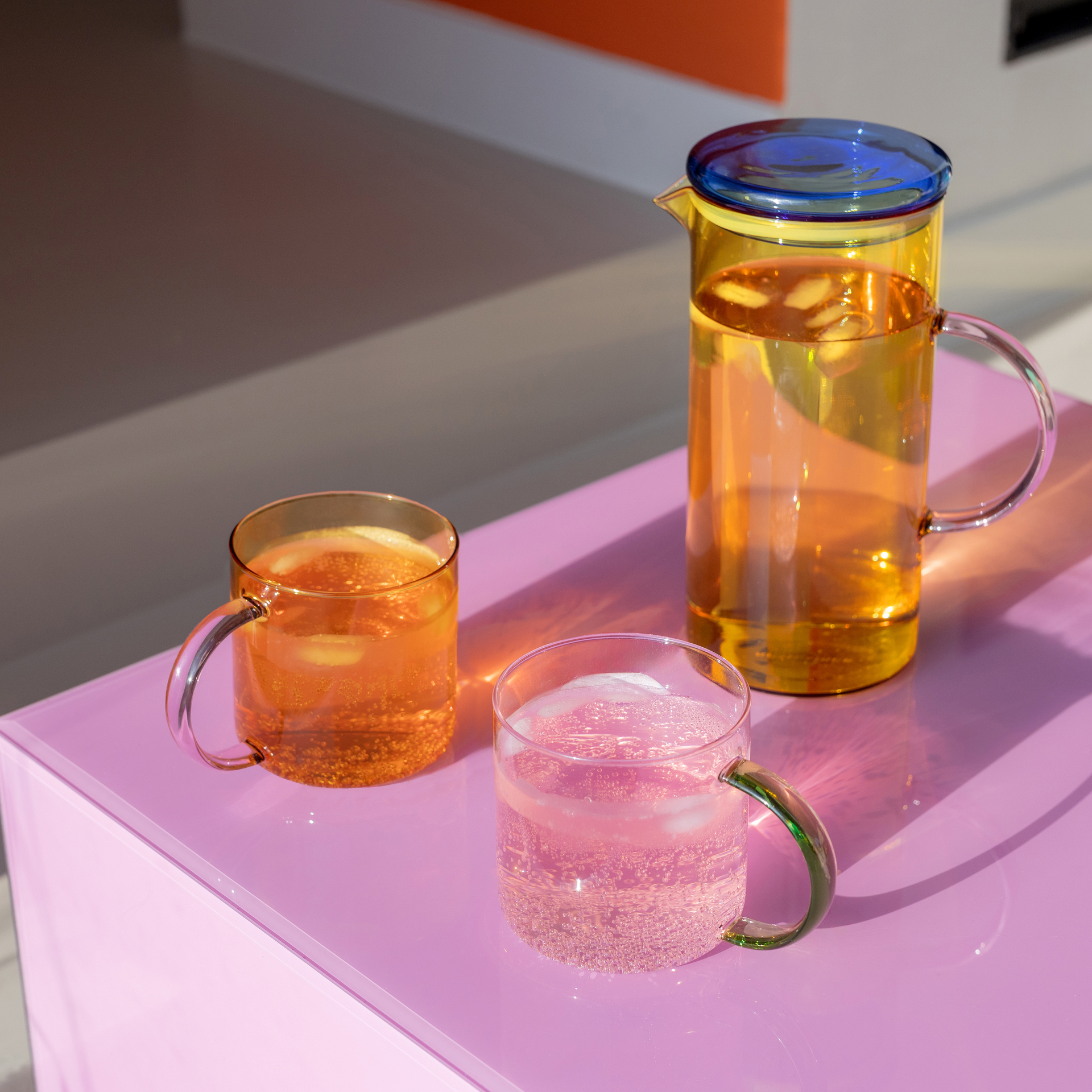 Gelbe Glaskanne mit blauem Deckel und zwei Gläser mit Getränken auf einem pinken Tisch.