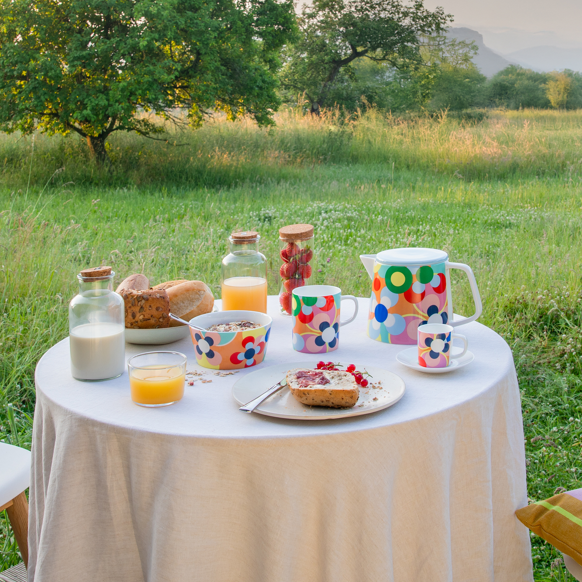 Ontbijttafel op het platteland: wit tafelkleed, kleurrijke kopjes/kannen, brood, muesli, sap, melk en aardbeien. Weiland, boom en bergen op de achtergrond.