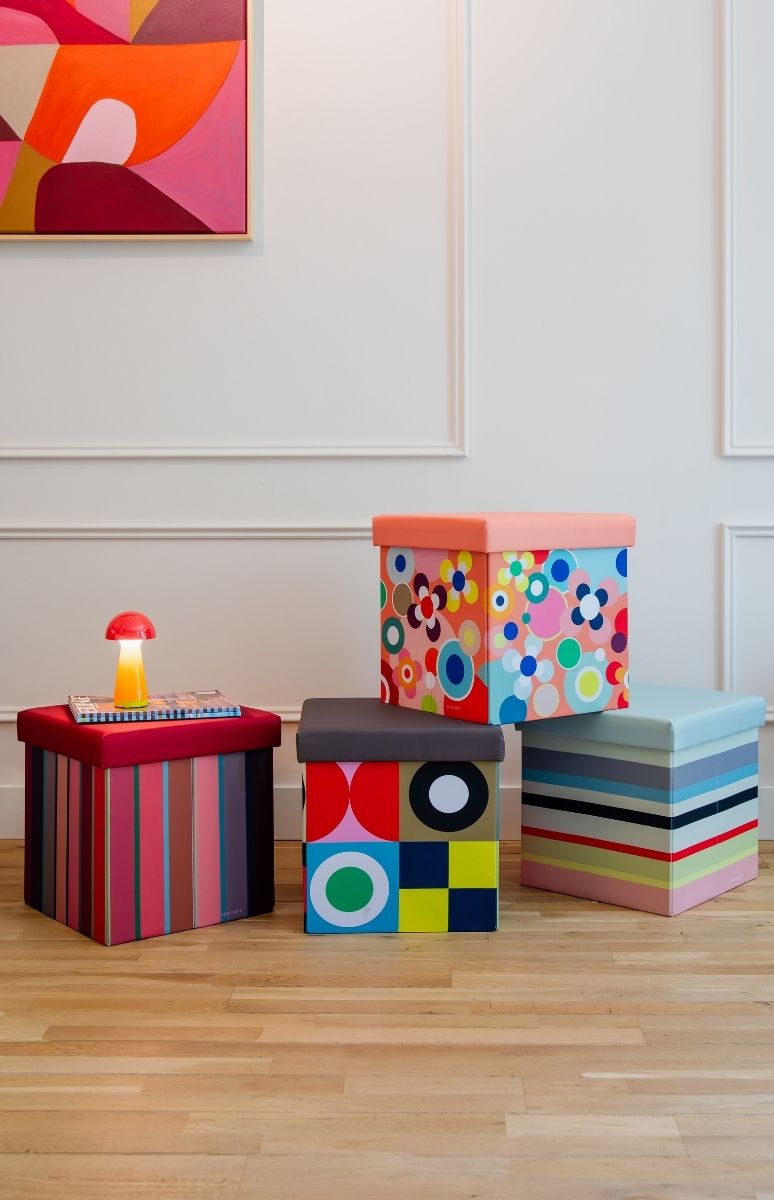 Vier bunte Aufbewahrungshocker mit verschiedenen Mustern stehen auf Holzboden: links rot gestreift mit pilzförmiger Lampe, mittig geometrisch, hinten floral gepunktet, rechts hellblau gestreift. Abstrakte Malerei an weißer Wand.