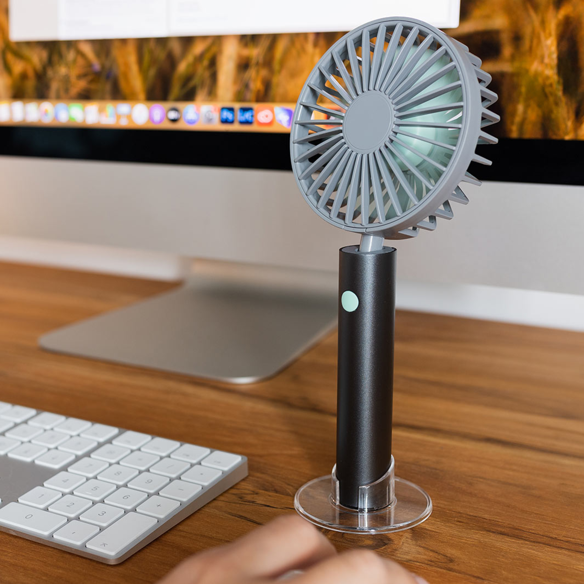 Un ventilateur Flow de couleur graphite avec un pied transparent est posé sur un bureau, à côté d'un clavier et d'un écran.