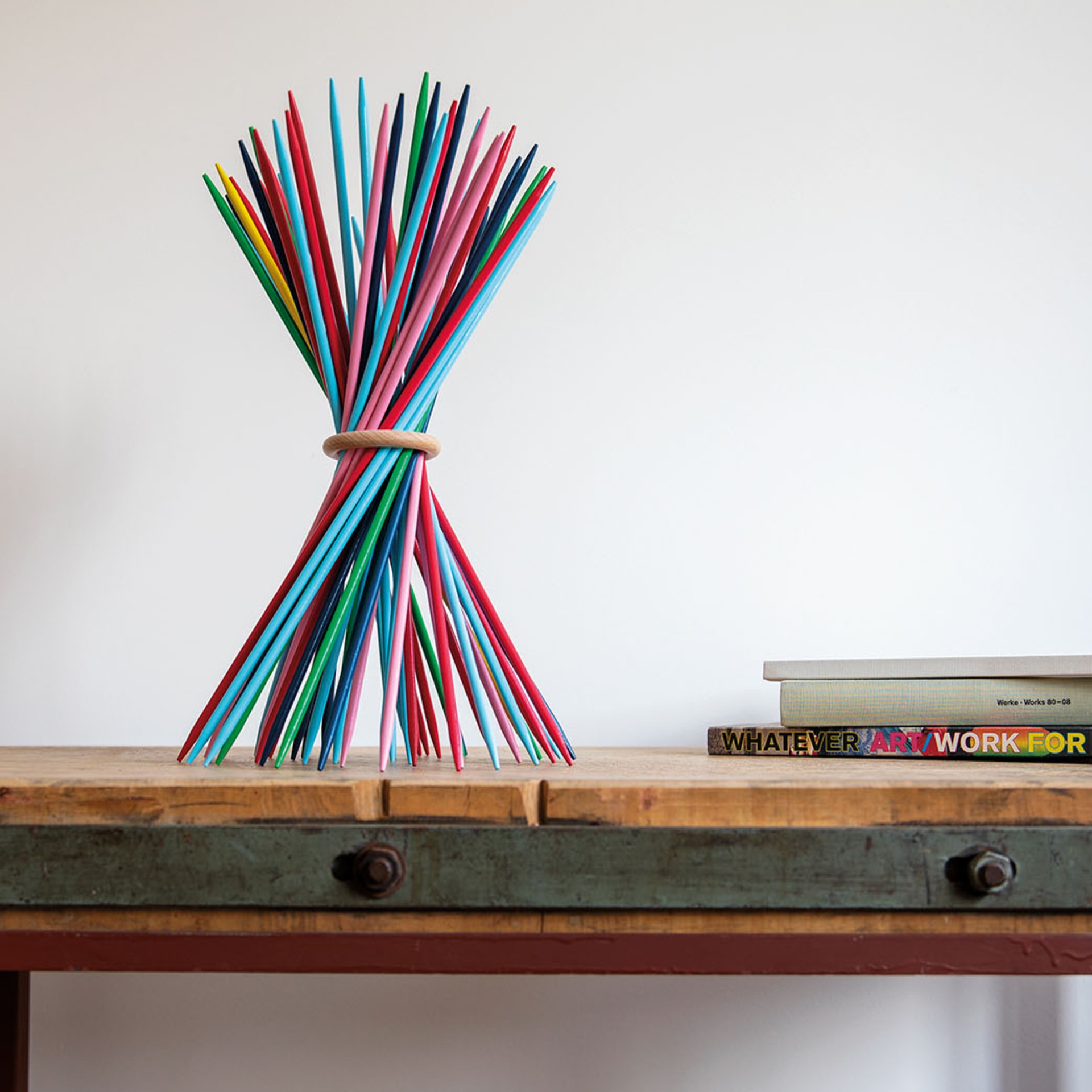 Een bundel kleurrijke houten stokjes, bijeengehouden door een houten ring, staat op een tafel. Op de achtergrond liggen boeken opgestapeld. De houten stokjes van Stickado hebben verschillende kleuren.
