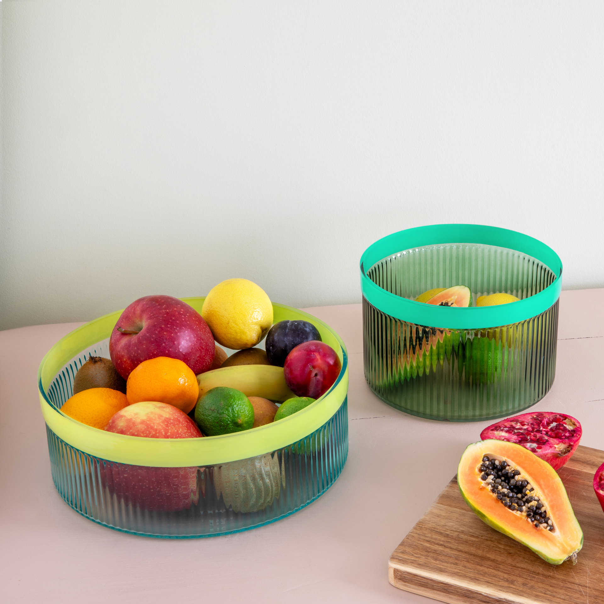 Twee glazen schalen, één gevuld met groen fruit zoals appels, citroenen en kiwi's. De andere schaal is leeg en ernaast liggen papaja's en granaatappels op een houten plank.
