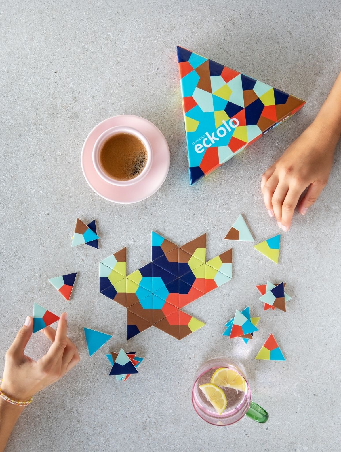 Draufsicht auf zwei Hände, die ein "eckolo" Geometrie-Puzzle auf hellgrauer Fläche legen. Bunte Dreiecke in Braun, Blau, Türkis, Gelb und Rot formen teils eine große Figur, teils liegen sie lose. Kaffeetasse und Zitronenwasser.