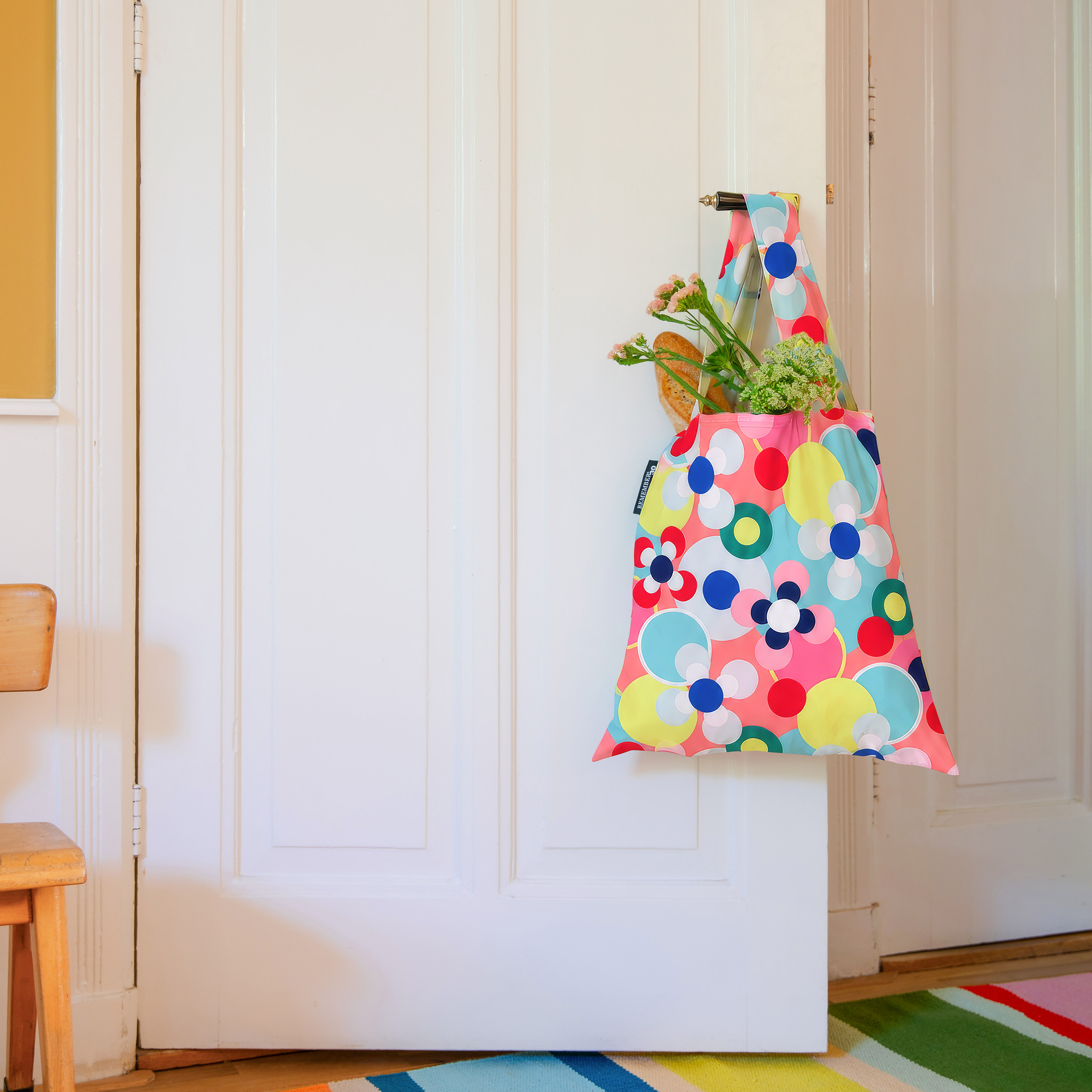 Eine bunte Tragetasche hängt an einem Türgriff einer weißen, leicht geöffneten Tür. Sie ist mit Kreisen und Blumen in Pink, Rot, Blau, Grün, Gelb und Weiß gemustert. Darin stecken ein Baguette und rosa-grüne Blumen.