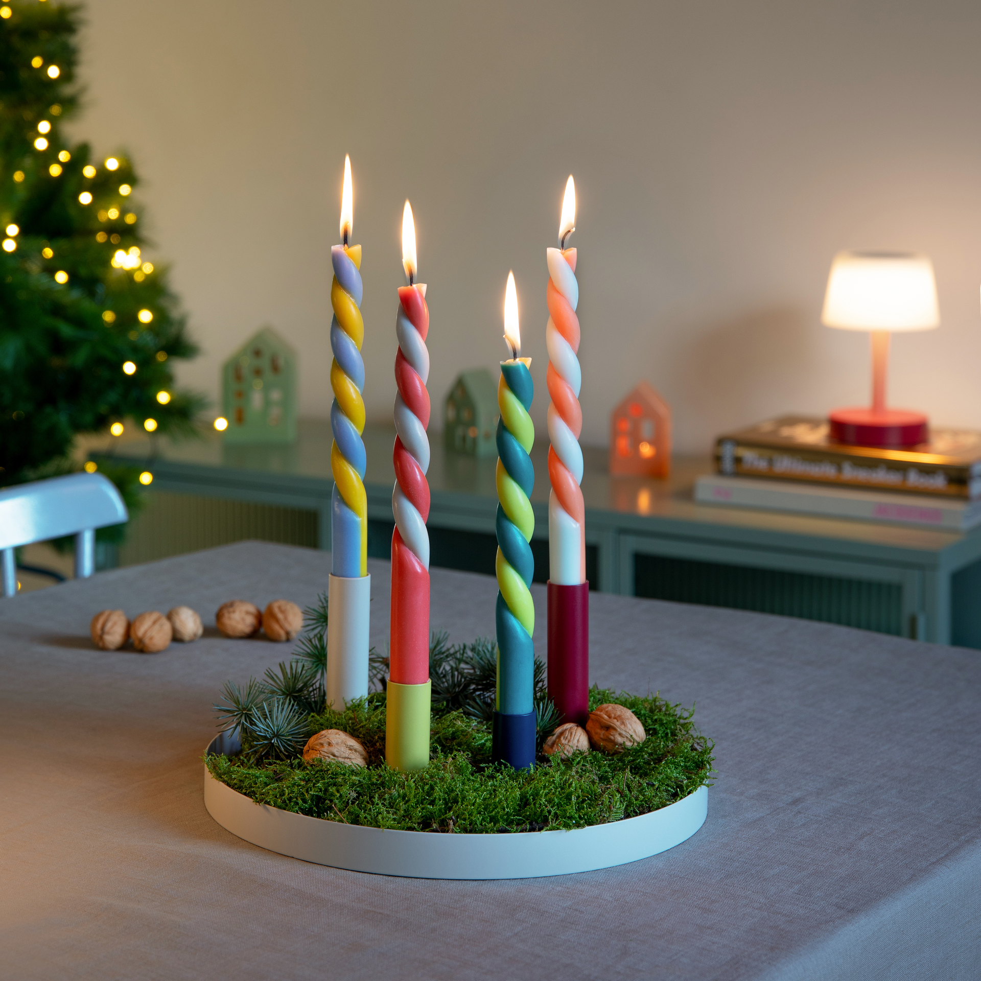 Cuatro velas cilíndricas de colores sobre un lecho de musgo en una bandeja blanca para velas. Las velas están encendidas, con un árbol de Navidad iluminado al fondo.