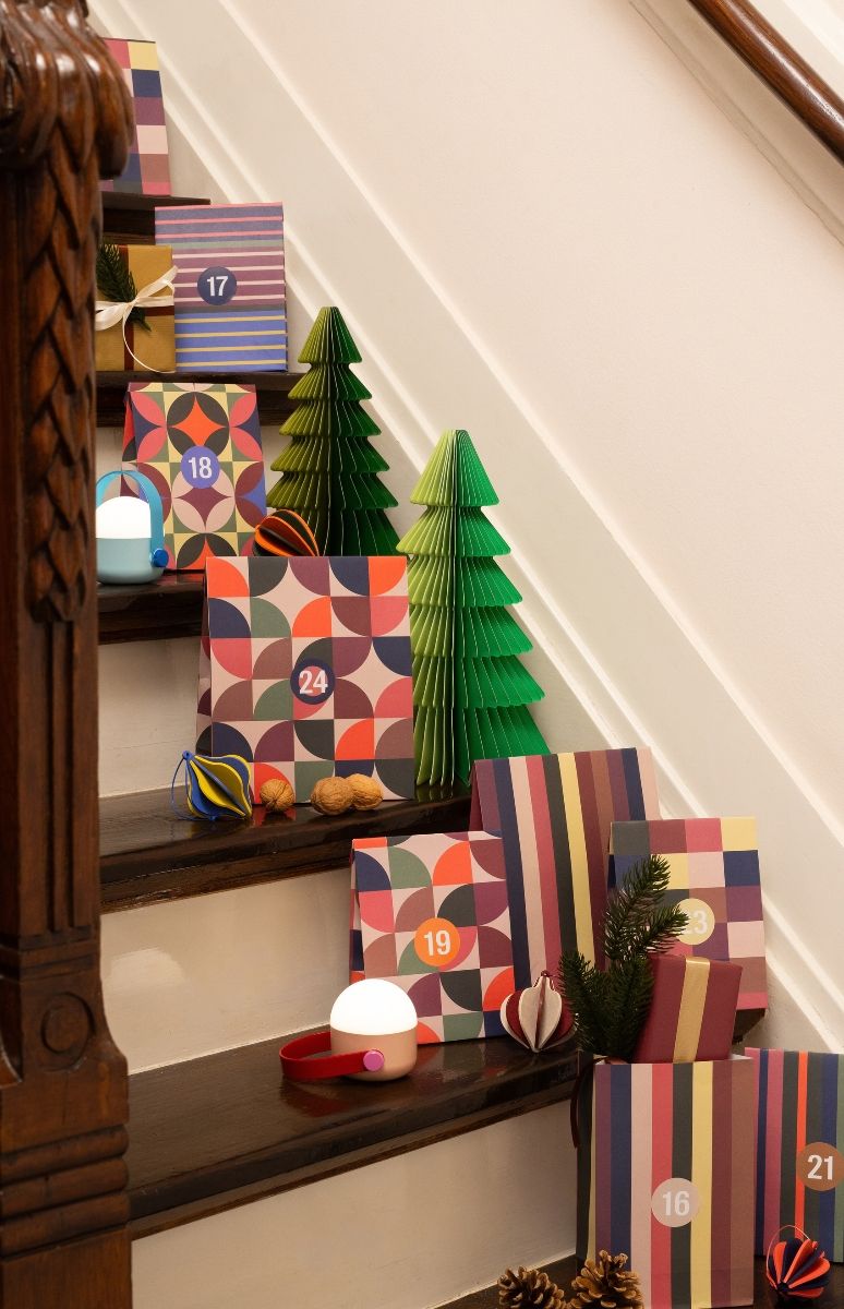 Geschmücktes Treppenhaus mit Adventskalender aus bunten Geschenkpaketen. Auf dunklen Holzstufen liegen Geschenke mit geometrischen Mustern und Streifen, nummeriert 16–24. Dazu grüne Papierbäume, Licht, Walnüsse, Deko.