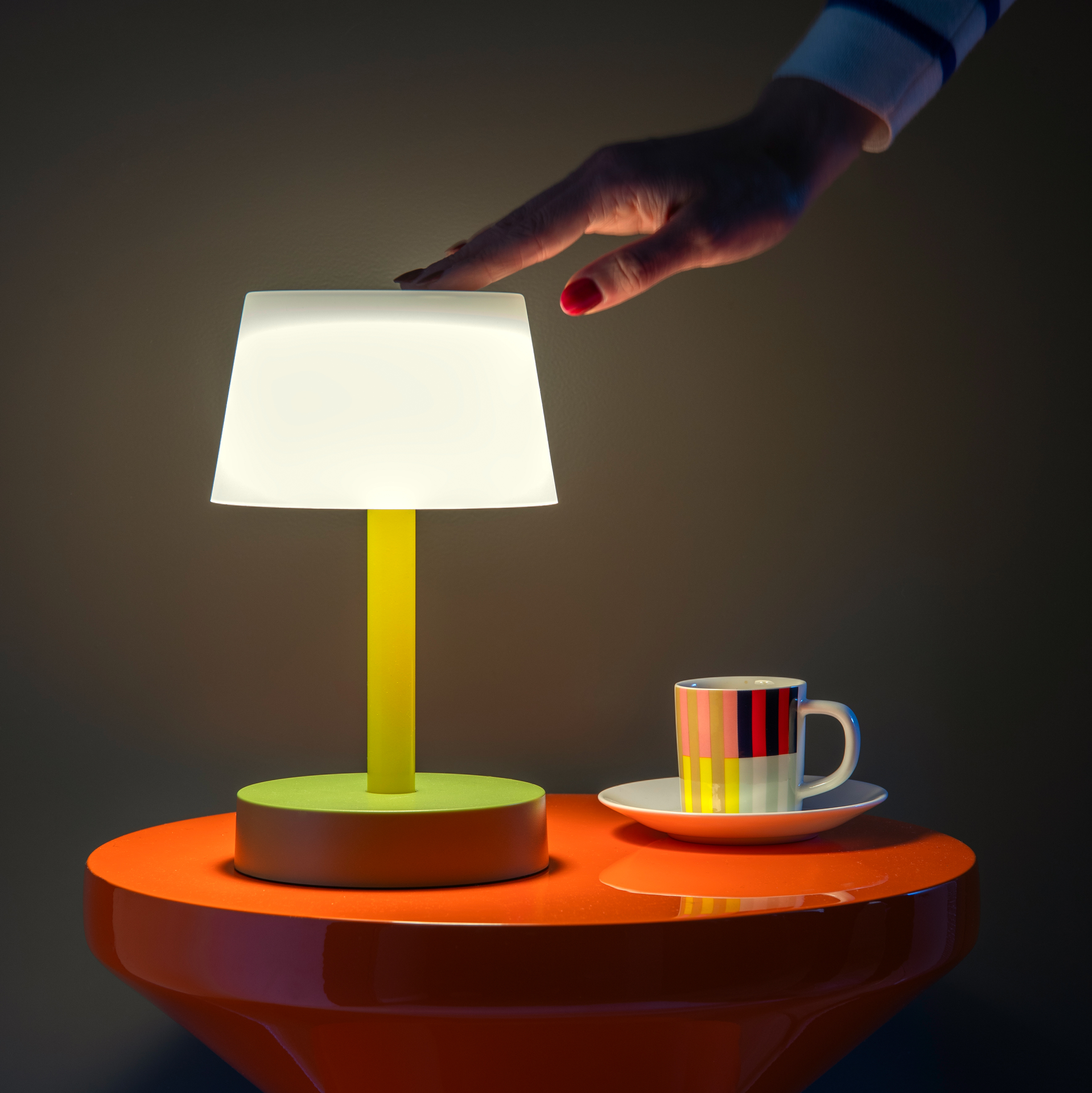 Une main touche l'abat-jour de la lampe de table Fritz 'flora'. La lampe, avec une tige jaune et un pied vert, est posée sur une table orange. Une tasse est posée à côté.