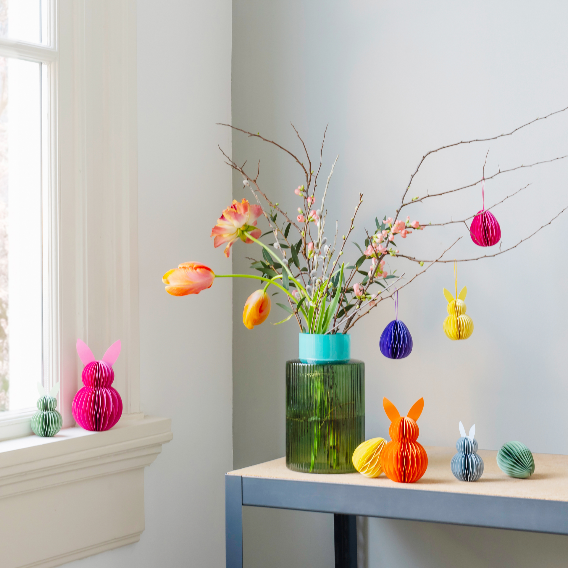 Decorative Easter eggs and paper bunny tree decorations stand next to a vase with branches and flowers on a table. Some of the decorations are hanging from the branches.