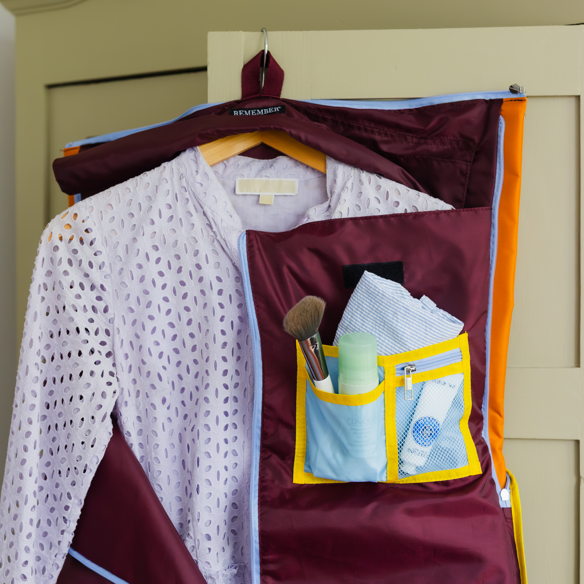 Aufgeklappte Reisetasche Lennard in tiefem Bordeauxrot hängt an einem Haken. Ein hellviolettes, gelochtes Kleidungsstück hängt knitterfrei auf Holzbügel im Hauptfach. Rechts Organizer-Fächer mit Pinsel, Deo und Kosmetiktube.
