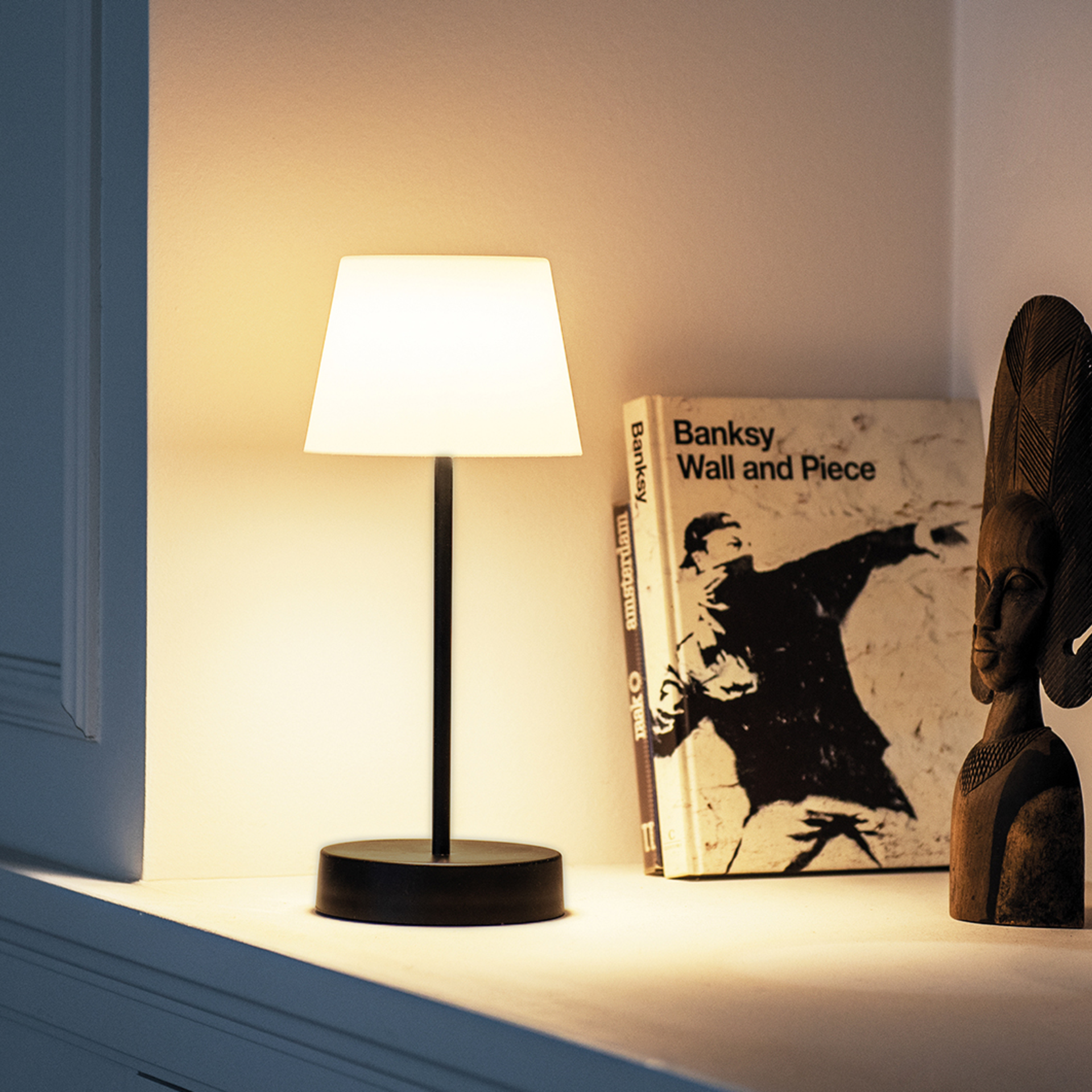 The Oscar 'pure' table lamp stands on a ledge. The lamp has a black base and stem, the lampshade is light-coloured. A book and a wooden figure stand next to it.