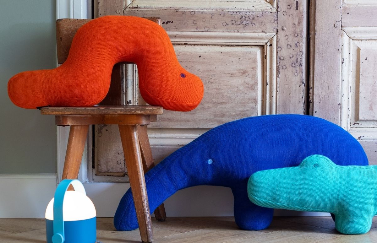 Orange Wurm-Kissen auf Holzschemel. Daneben auf hellem Boden ein dunkelblauer Kuschelwal und ein türkisfarbenes Krokodil-Kissen. Vorn eine blau-weiße Laterne mit Licht.