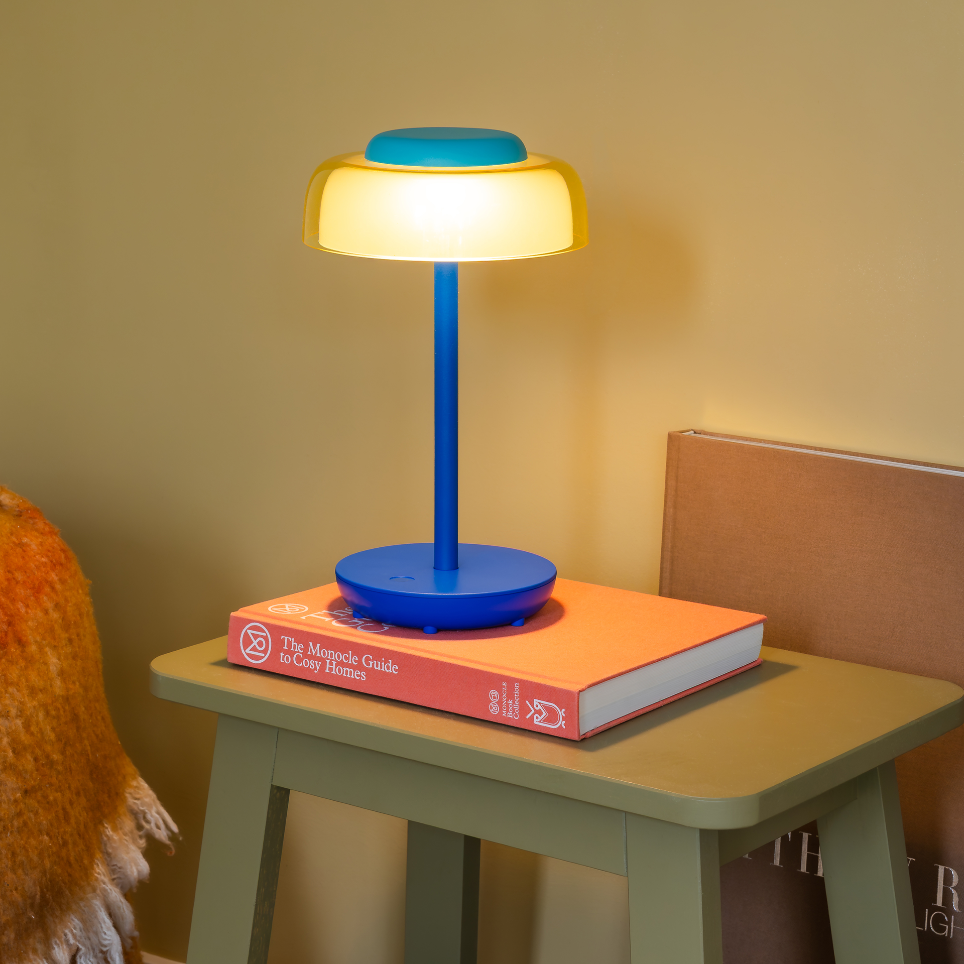 Moderne, draadloze tafellamp Juno met een slanke, donkerblauwe voet en een ronde basis. Geel-transparante lampenkap met een heldere koepel. Het platte, turkooiskleurige aanraakoppervlak en het aan/uit-symbool zorgen voor een intuïtieve bediening.