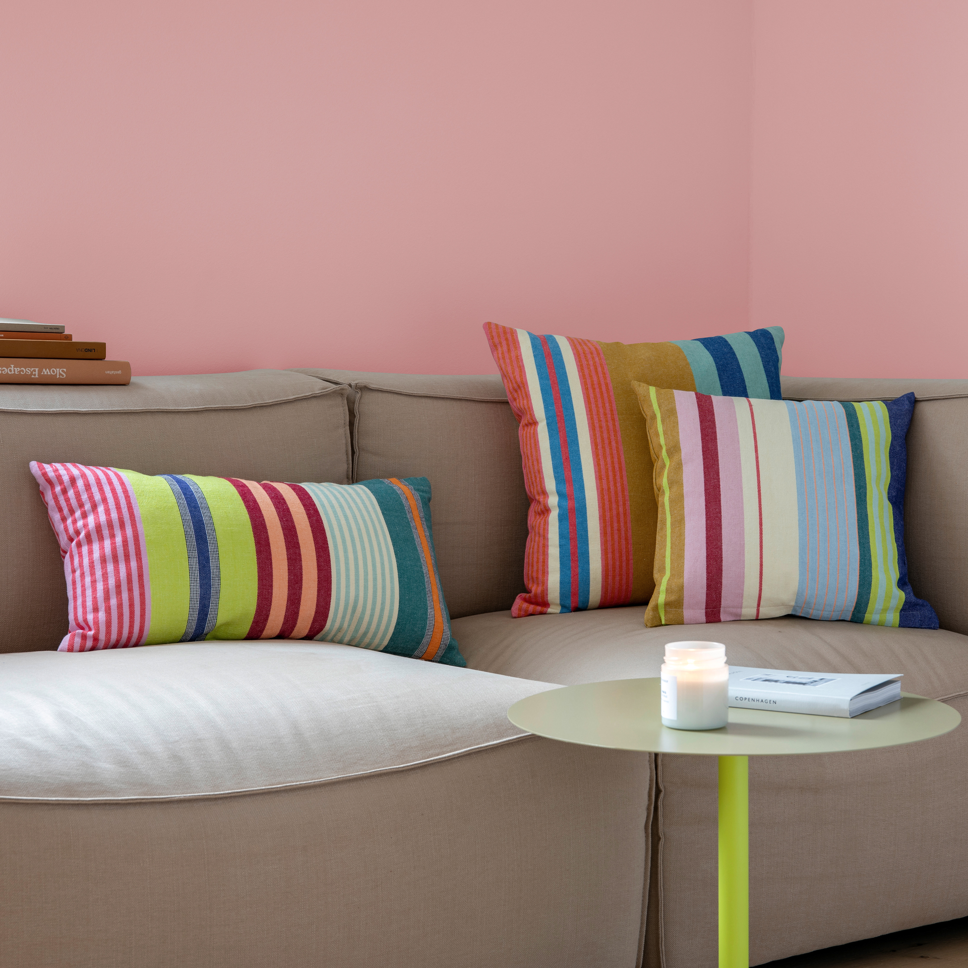 Beige bank met drie gestreepte katoenen kussens 'Barbados' in felle kleuren. Een klein, rond tafeltje met een boek en een kaars staat ervoor, de muur is roze.