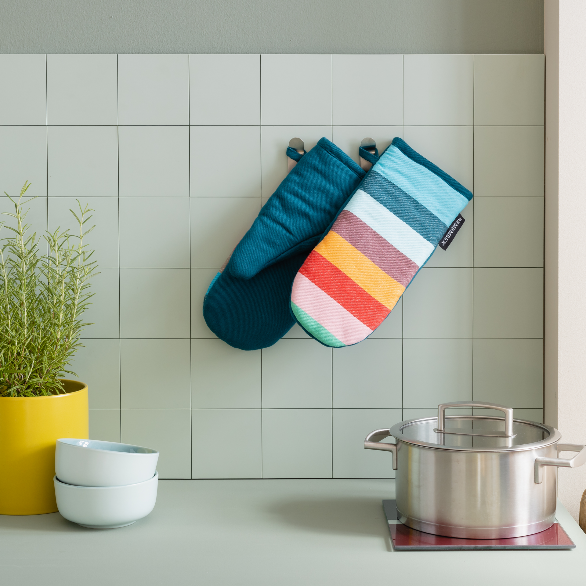 Zwei Ofenhandschuhe im Set, gestreift in Farben wie Blau, Rosa und Orange, liegen übereinander. Die Handschuhe haben eine Schlaufe zum Aufhängen.