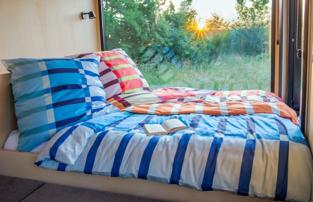 Ein gemütliches Bett mit bunten blau-weiß karierten und orange-rot gestreiften Decken steht vor einem großen Fenster mit Blick auf eine grüne Landschaft bei Sonnenauf- oder -untergang. Ein offenes Buch liegt bereit.