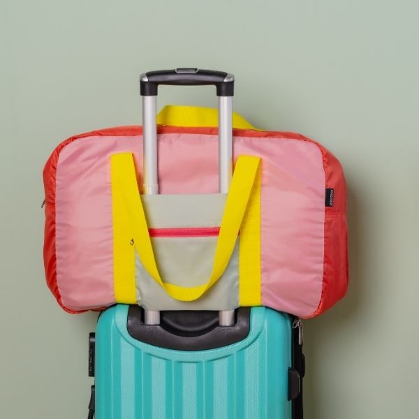 Eine farbenfrohe Reisetasche in Pink und Rot mit gelben Tragegriffen, die fest auf dem ausziehbaren Griff eines türkisfarbenen Hartschalenkoffers sitzt. Der Koffer steht vor einer hellgrünen Wand, bereit zur Abreise.