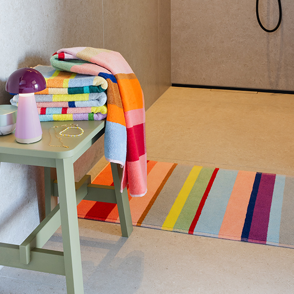 Badezimmer: hellgrüner Hocker mit Stapel bunter Handtücher, lila Pilzlampe und goldenem Schmuck. Ein gestreiftes Handtuch hängt darüber. Auf hellem Fliesenboden liegt ein langer, diagonal gestreifter Badteppich. Duschkabine im Hintergrund.