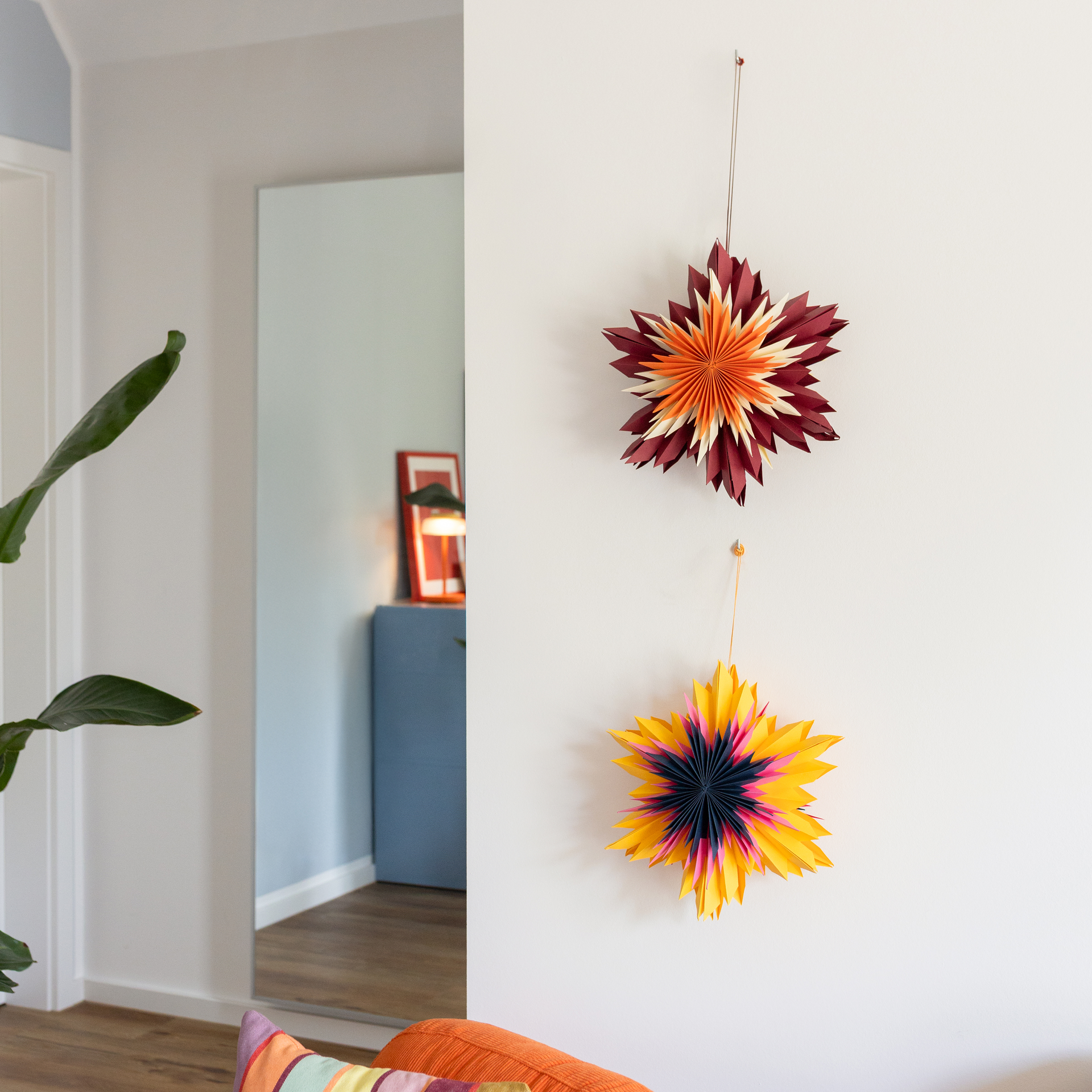 Dos estrellas decorativas de papel del conjunto Nova cuelgan de una pared blanca. La superior tiene estampado en rojo burdeos, naranja y crema; abajo brilla una estrella amarilla, rosa y azul oscuro. Ambas bien plegadas.