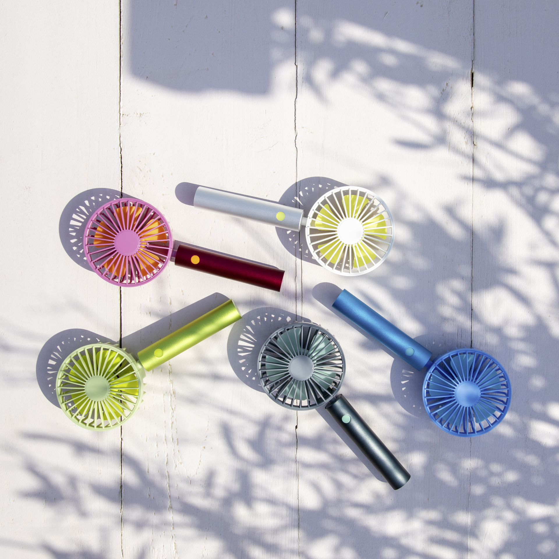 Cinq ventilateurs à accu colorés et rechargeables sur une table en bois clair. Les couleurs sont le rose, l'argent, le rouge foncé, le vert et le bleu. Ombre de feuilles en arrière-plan.