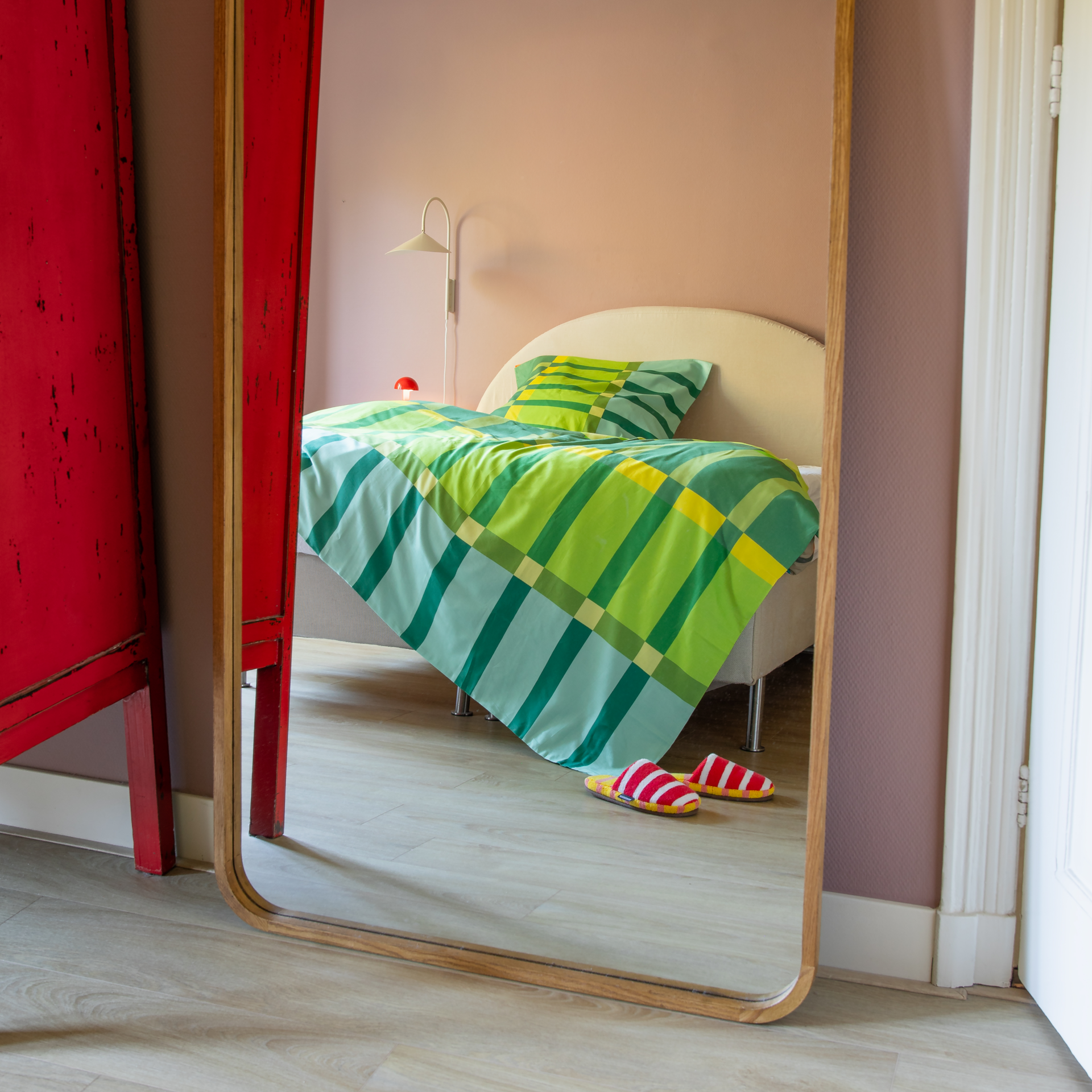 Spiegelung eines Bettes mit gestreifter Vito-Bettwäsche in Grün- und Gelbtönen. Decke und Kissen zeigen ein modernes Muster aus seidenweichem Mako-Satin. Rote Hausschuhe auf hellem Holzboden, dazu Wandleuchte und rote Lampe.
