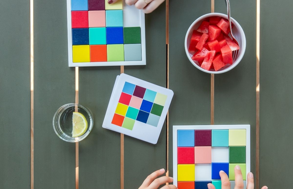 Zwei Hände ordnen bunte Quadratblöcke in zwei weißen Tabletts. Daneben Kartenstapel, Glas Wasser mit Zitrone und eine Schale Wassermelone auf einem grünen Tisch.