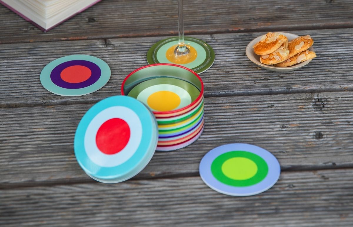 Kleurrijke, ronde onderzetters met concentrische cirkels liggen op een rustieke houten tafel. Sommige zijn gestapeld in een cilindervormig blik met horizontale strepen. Er staat een wijnglas op, met daarnaast een schaal met amandelkoekjes.