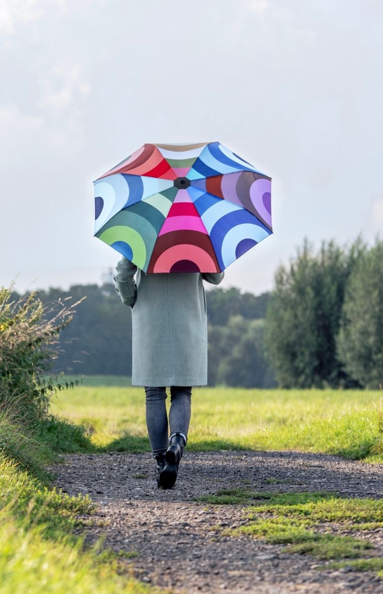 Eine Person geht von hinten auf einem Schotterweg durch eine grüne Landschaft. Sie trägt einen langen mintgrünen Mantel und hält einen großen, bunten Regenschirm mit konzentrischen Kreisen in Rot, Blau, Grün und Rosa.