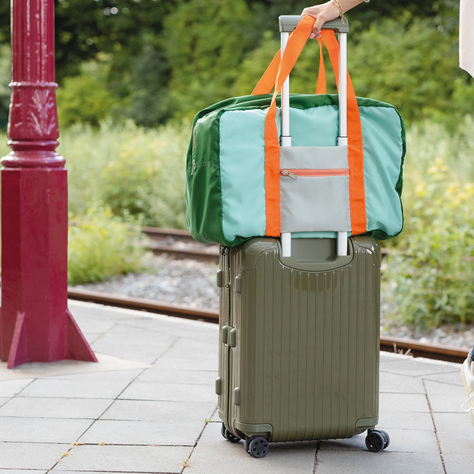 Una bolsa de viaje verde «Max» con asas naranjas descansa sobre una maleta con ruedas de color verde oliva. Ambas se encuentran en un andén, con un fondo verde.