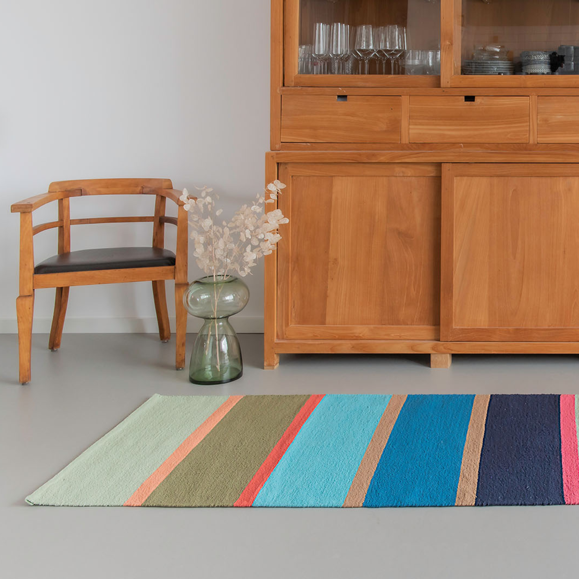 Un tapis de sol 'Costa' à rayures multicolores est posé devant une armoire en bois et une chaise. Un vase avec des fleurs séchées est posé à côté.
