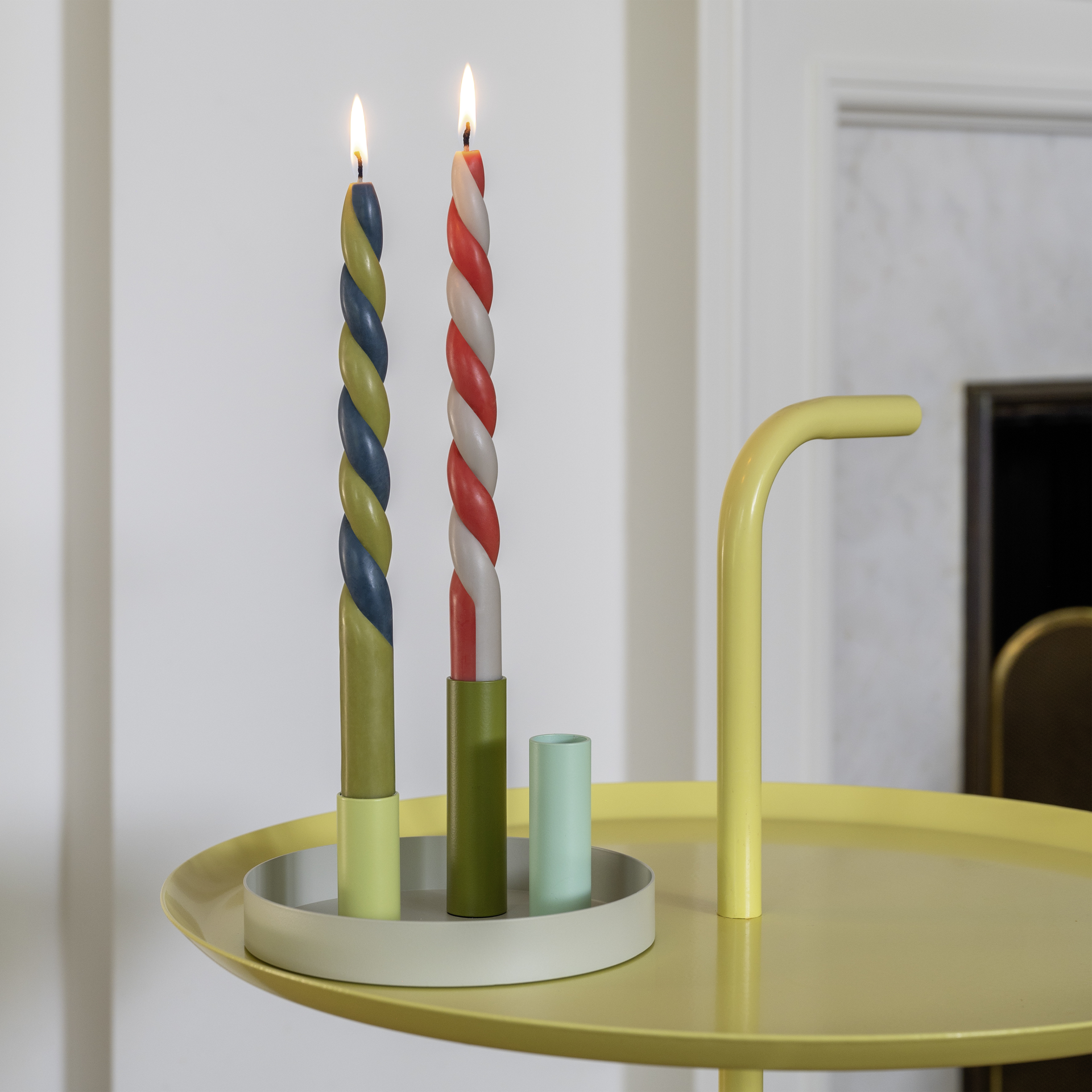 Portavelas magnético «Luna Mini» con tres portavelas de colores y dos velas rizadas encendidas sobre una mesa amarilla.