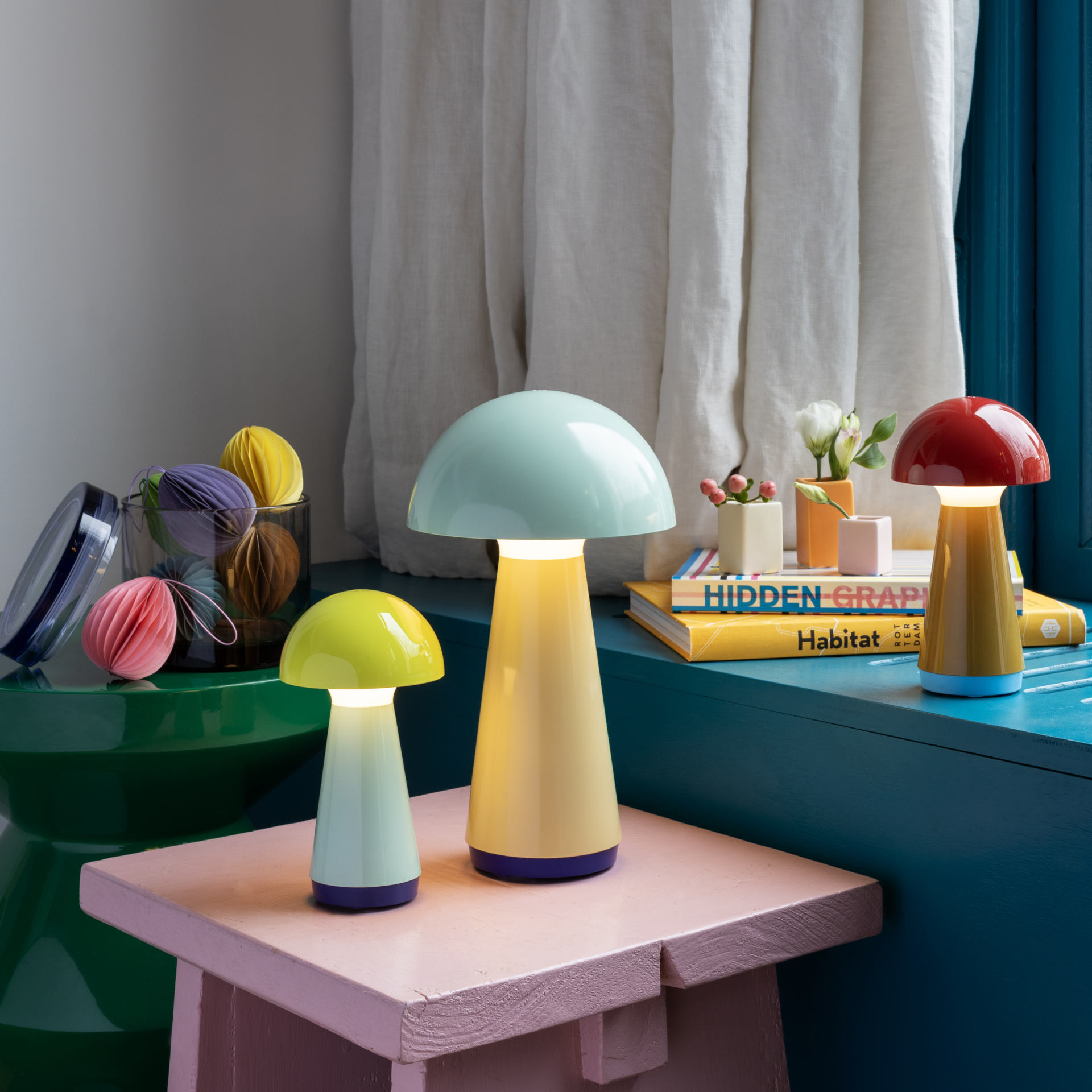 Tres lámparas de mesa «Bob» en color menta, amarillo y rojo descansan sobre mesas y libros. Tienen forma de seta y emiten una luz blanca cálida. Al fondo, una ventana con cortinas.