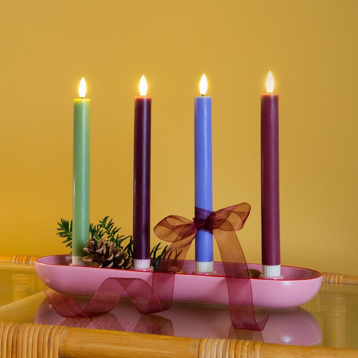 Vier brennende Kerzen in Grün, Violett, Blau und Burgunder stehen in einer rosa Adventsschale. Sie sind dekoriert mit Tannengrün, einem Zapfen und einem roten Schleifenband. Das Arrangement steht auf einer spiegelnden Oberfläche vor einer gelben Wand.