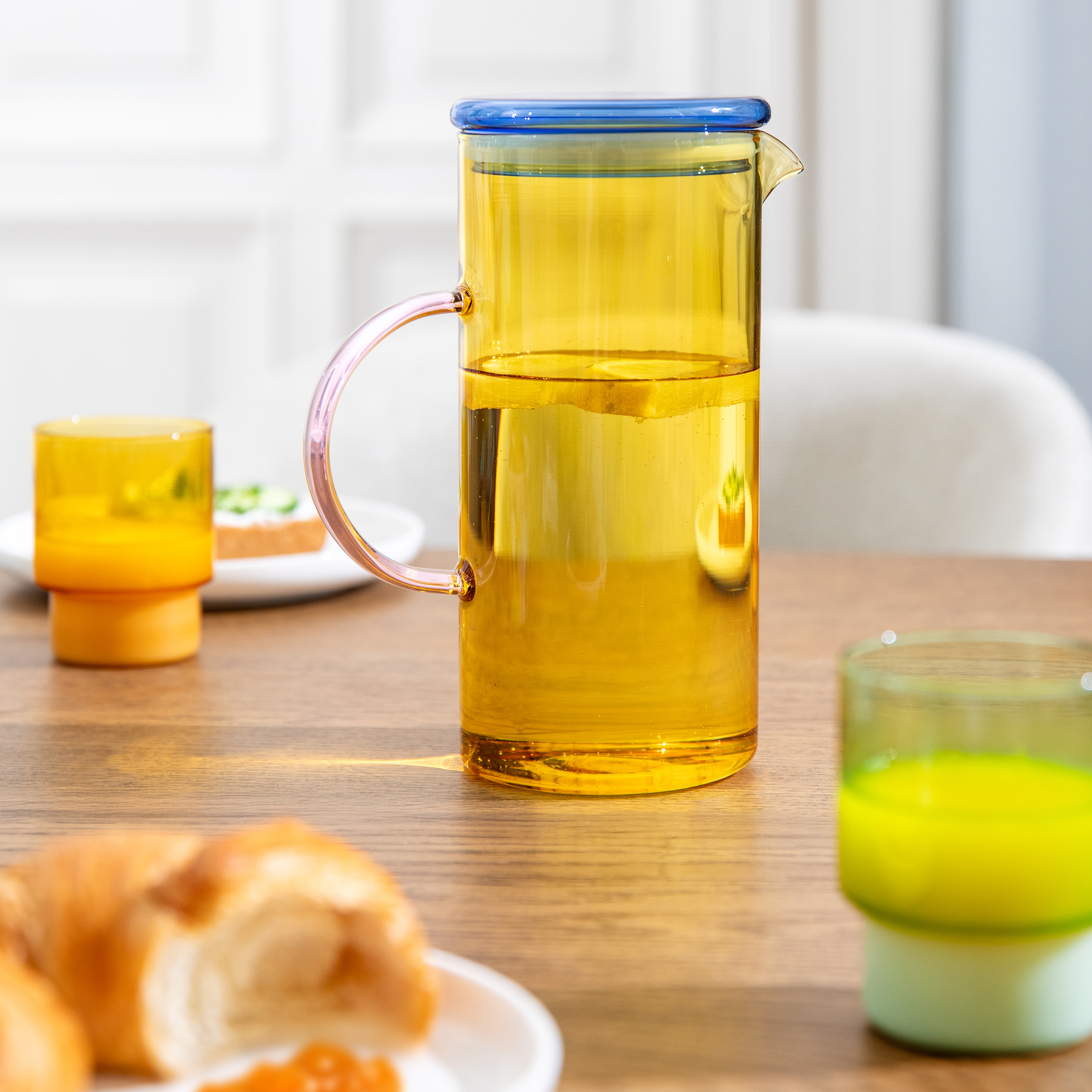 Ein Tisch mit Frühstück. Im Fokus steht eine gelbe Glaskaraffe mit blauem Deckel und rosa Henkel, gefüllt mit Flüssigkeit und Zitronenscheiben. Dazu Croissant, Marmelade und Getränke.