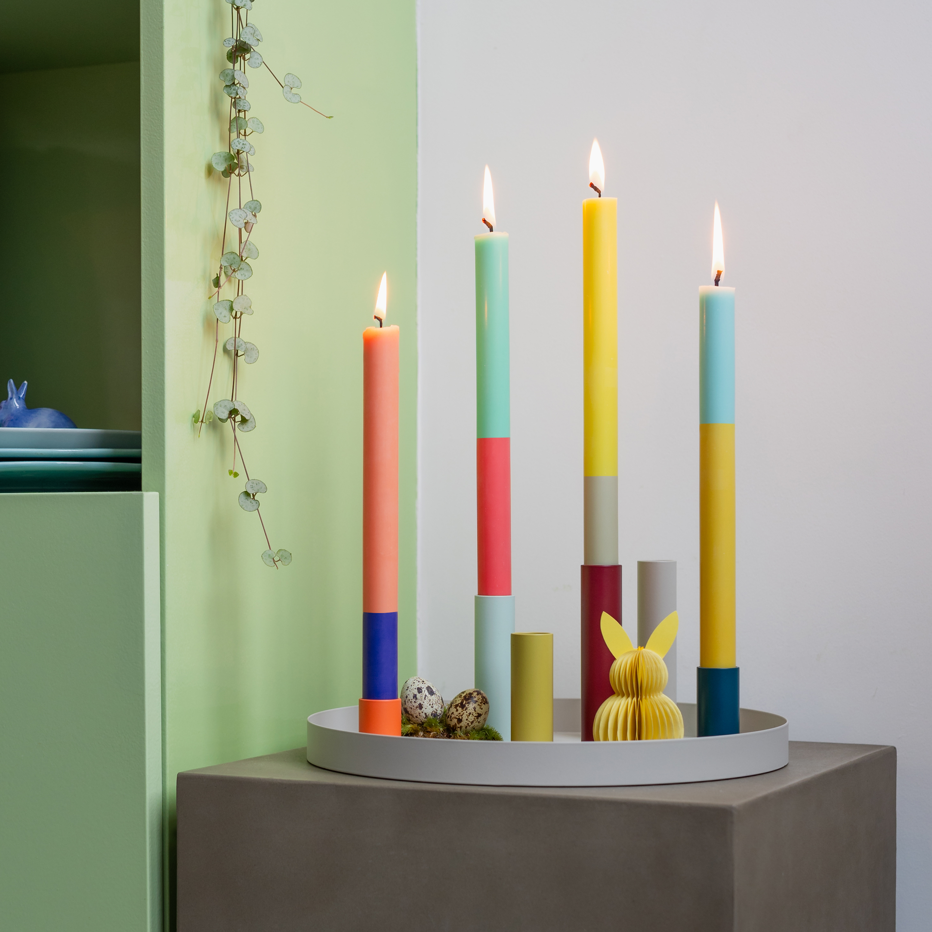 Candele a bastoncino "Ibiza" colorate e ardenti, con un design a blocchi di colore su un vassoio rotondo, completate da uova decorative e un coniglietto di carta.