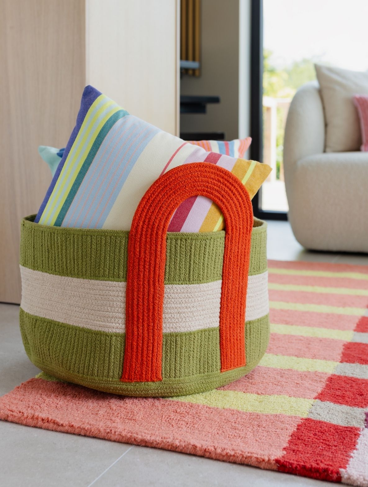 Olivgrüner runder Korb mit beigen Streifen auf einem mehrfarbigen Teppich. Darin mehrere Kissen mit bunten Streifen in Gelb, Blau, Rosa und Grün. Orangefarbener Bogenhenkel. Im Hintergrund heller Wohnbereich mit Sessel und Fenster.