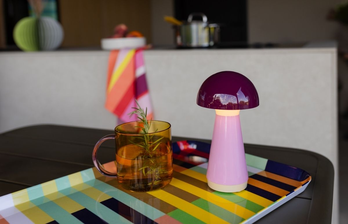 Bunte Szene: orangefarbener Becher mit Getränk, Orangenscheibe und Rosmarin auf gestreiftem Tablett. Daneben eine eingeschaltete pilzförmige Lampe mit lila Kopf und pinkem Stiel. Hintergrund: Küche, gestreifte Decke.