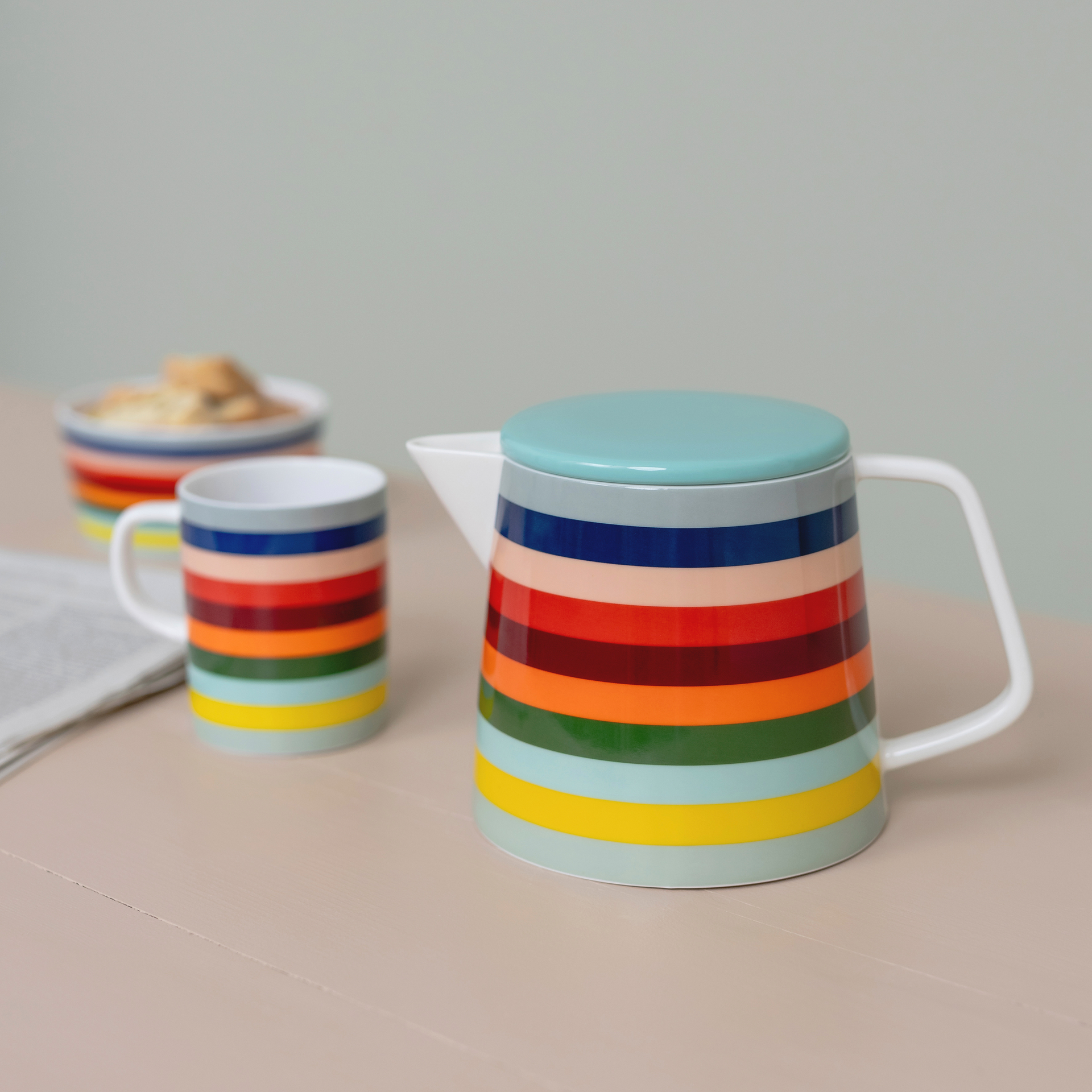 Teiera "Lorenzo" con coperchio turchese e strisce colorate, accanto a una tazza con lo stesso disegno e a una ciotola con biscotti.
