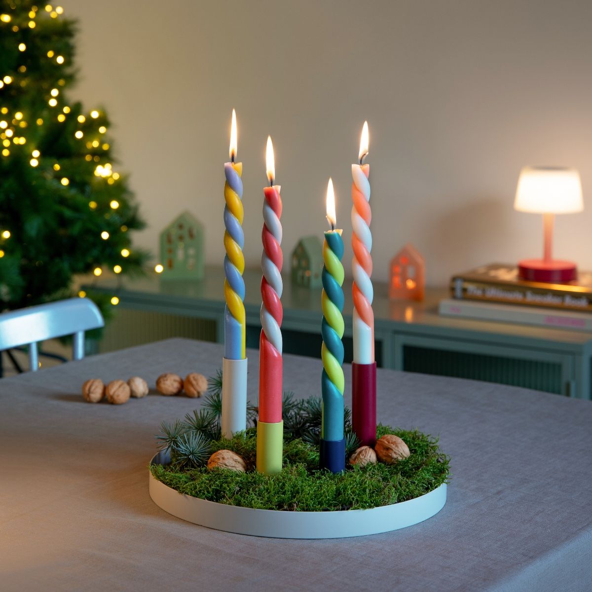 Festlicher Adventskranz mit vier bunten, spiralförmigen Kerzen, die hell leuchten. Auf einem Kranz aus grünem Moos mit Tannenzweigen und Walnüssen. Im Hintergrund: beleuchteter Weihnachtsbaum und Sideboard mit Lichthäusern.