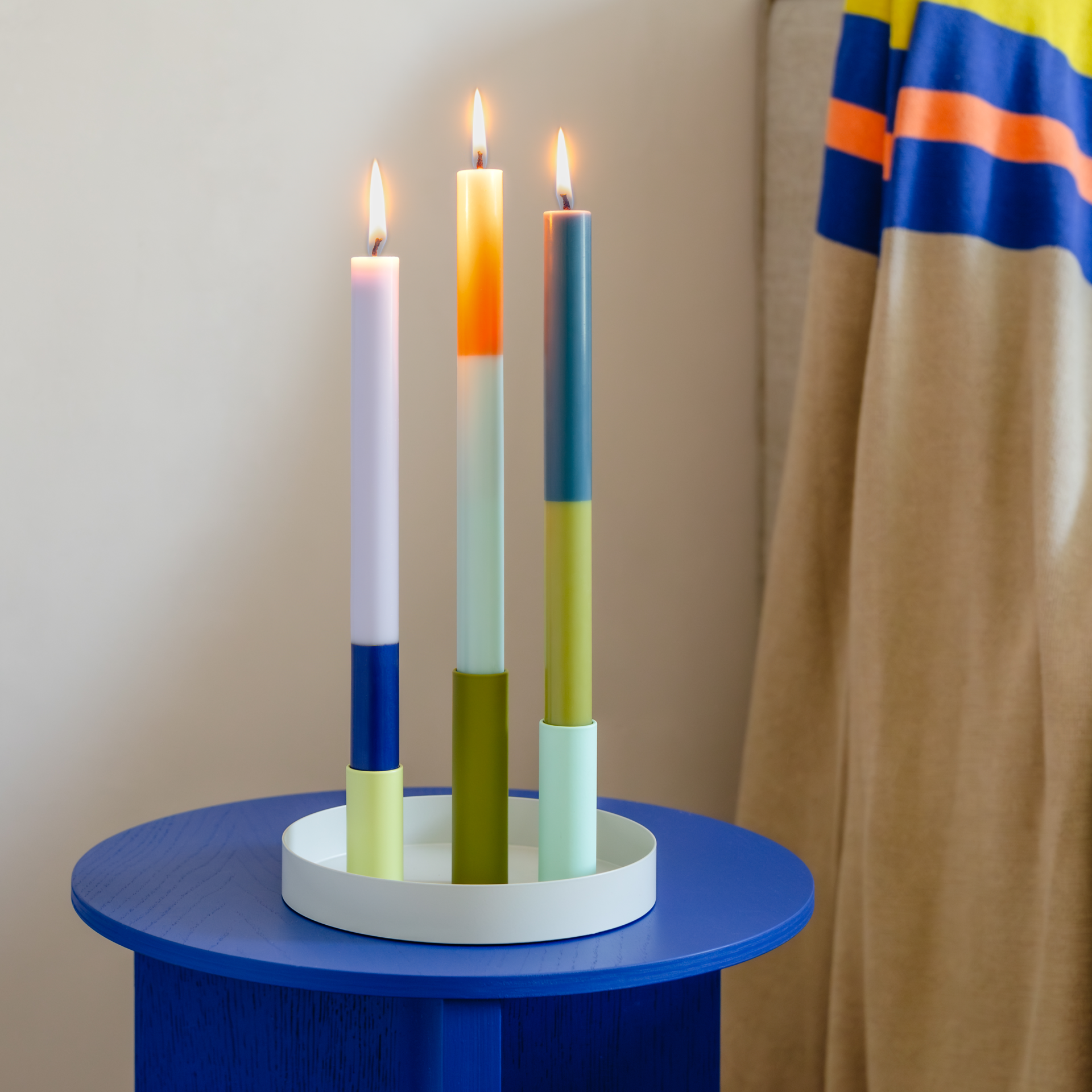 Tres velas cilíndricas de colores del juego de candelabros «Luna Mini» descansan sobre una mesa azul. Los candelabros combinan en color con la mitad inferior de las velas. Todas las velas están encendidas.
