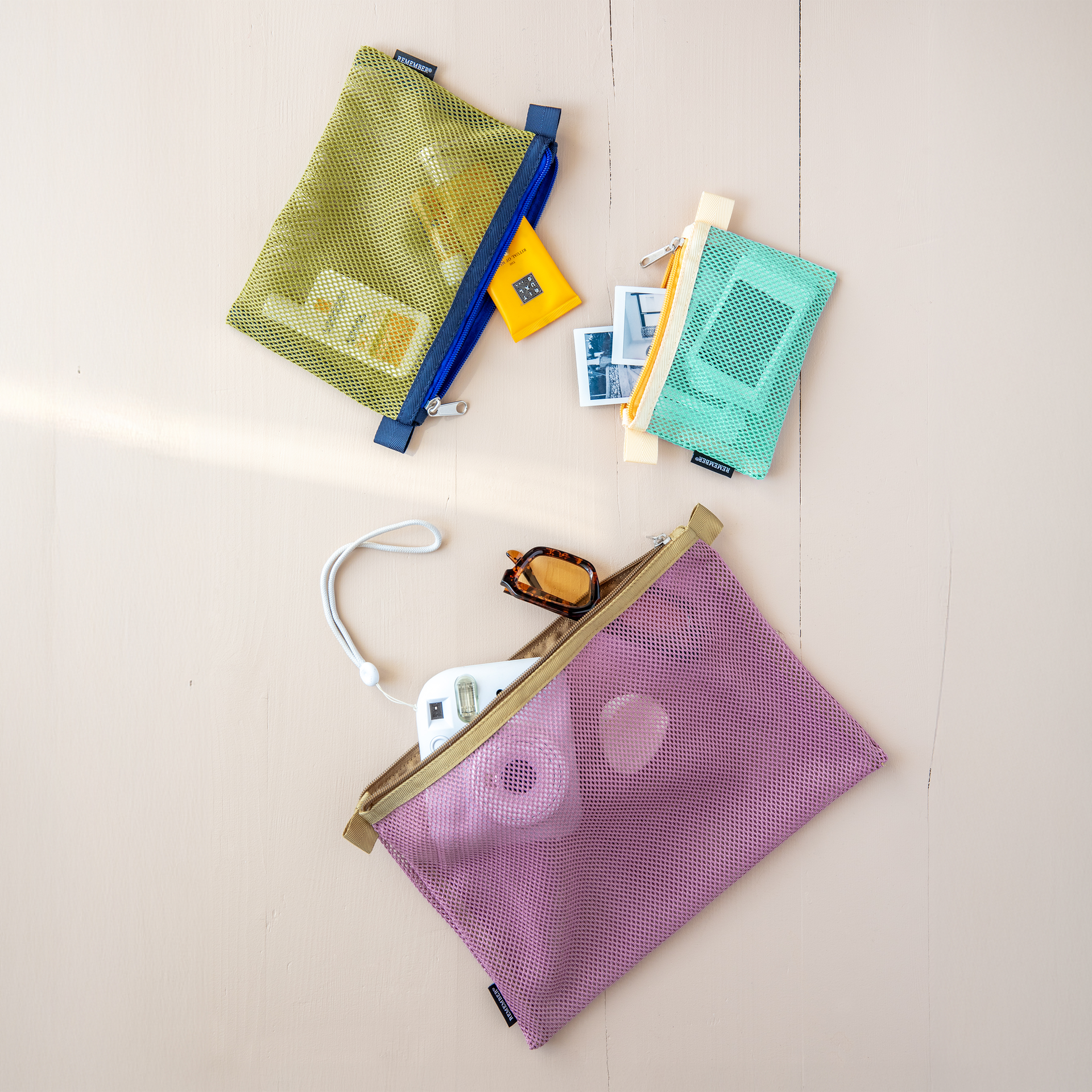 Three coloured mesh bag sets, open on one surface. The colours are green, turquoise and purple. The bags contain cosmetics, sunglasses and a camera.